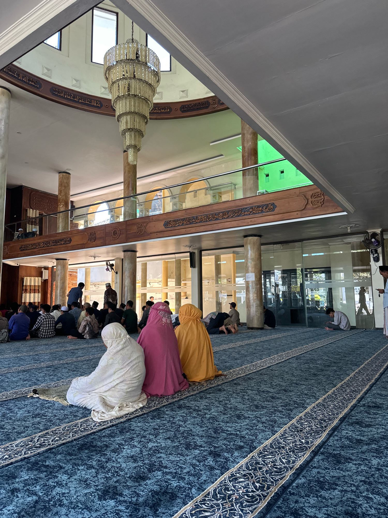 The image depicts the interior of a mosque, likely the Great Mosque of Malang (Masjid Agung Jami' Malang) in Malang, Indonesia, based on the architectural style and typical use of such spaces. In the foreground, several individuals, predominantly women wearing hijabs and modest clothing in shades of white, pink, and yellow, are seated on a blue carpeted floor, facing away from the camera. Some are positioned on prayer mats. Behind them, a group of men and women are also seated on the carpet, some in conversation or attending to prayer. A few men are standing, including one near a door and another near a fan mounted on a column.
The mosque's architecture features tall, cylindrical columns supporting an upper level with arched windows. A prominent, multi-tiered crystal chandelier hangs from the high ceiling, adorned with decorative Arabic calligraphy on the surrounding wooden beams. The overall environment is well-lit, with natural light filtering in from windows and internal lighting visible. The flooring is a patterned blue carpet, and the walls appear to be a neutral color with wooden decorative elements. The scene captures a moment of prayer or communal gathering within the mosque.