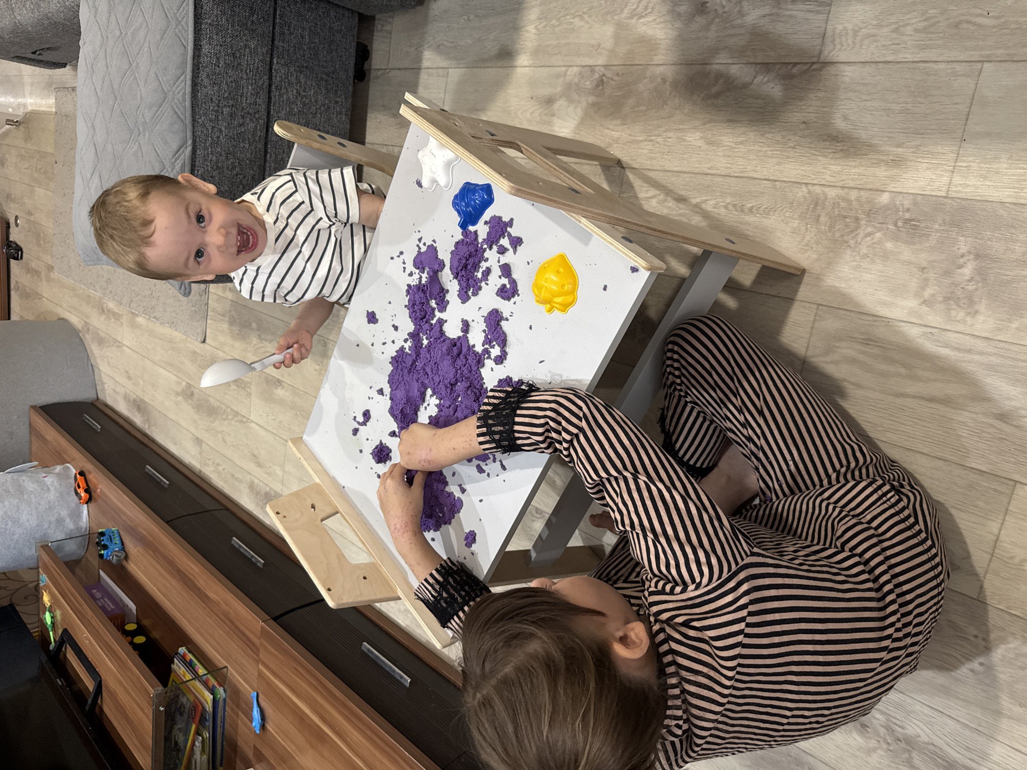 Two children are engaged in sensory play at a wooden activity table on a light-colored wood floor, likely indoors. One child, wearing a white and black striped shirt, is holding a spoon and looking at the camera with an open mouth. The other child, wearing a brown and black striped top and pants, is manipulating purple kinetic sand on the table. The table also has small containers of white, blue, and yellow play material. Behind the activity table is a brown cabinet, housing toys and books. The location is suspected to be in a residential area, possibly Rusanovo, Russia.