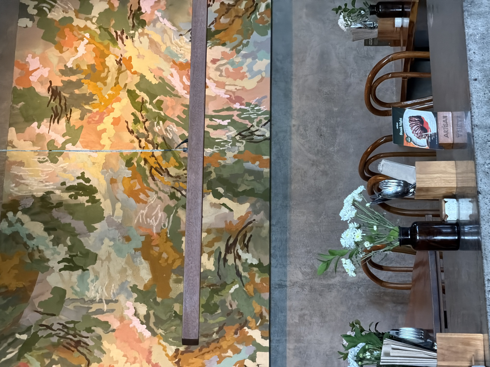 In Mengwi, Indonesia, this image captures a close-up view of a restaurant's dining area. A large, abstract mural with a blend of earthy tones, greens, and pops of orange and pink dominates the wall above a wooden light fixture. In the foreground, a dark polished table reflects the overhead lighting. On the table, several arrangements are visible: to the left, a metal napkin holder with folded napkins and a small vase with white flowers; in the center, a dark brown glass vase holds a bouquet of white flowers and green stems; and to the right, a small wooden stand holds cutlery. A menu board with an image of a steak and the text "ARTISAN Steak Night" is also displayed on the table. Behind the table, several curved wooden chairs are visible, suggesting a seating area for diners. The background wall appears to be textured concrete, adding to the modern, minimalist aesthetic of the establishment.