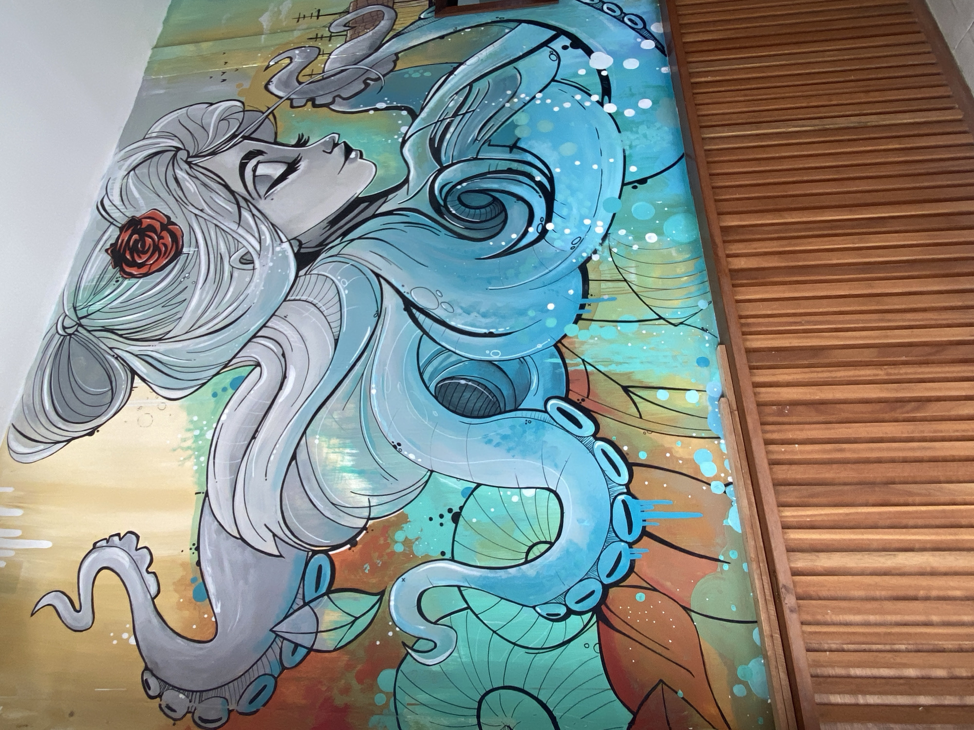 A detailed mural depicting a woman intertwined with an octopus is located on a wall in Baan Chaloklum, Thailand. The woman, with gray skin and gray hair adorned with a red rose, is looking upward. Her hair flows into the blue-toned tentacles of the octopus, which dominates the artwork. The background is a colorful mix of yellow, orange, and green, creating a surreal underwater scene. A wooden slatted structure is adjacent to the mural, partially obscuring the right side of the artwork.