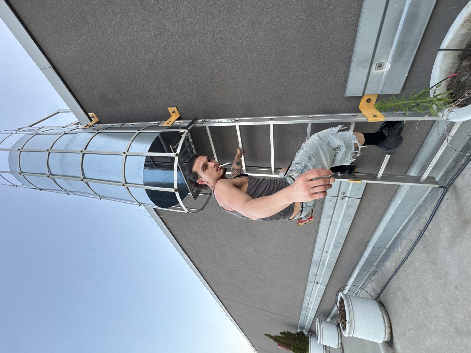 A young man with dark hair, wearing a grey tank top and ripped denim shorts, is sitting on a silver ladder outdoors. He is smoking a cigarette and looking towards the camera. His arms are bare, and he has tattoos on his left arm. The ladder is positioned against a large, grey, textured building. To the left of the man is a tall, cylindrical structure made of shiny, corrugated metal, possibly a silo or storage tank. The sky is a clear, pale blue, suggesting it is daytime. In the background, to the right of the ladder, a large white planter contains some green foliage. The overall scene appears to be a casual, possibly industrial or construction-related, outdoor setting. The image is taken from a low angle, looking upwards. There is no visible text in the image. The location context is Sofia, Bulgaria, but no specific landmarks are visible to confirm this.