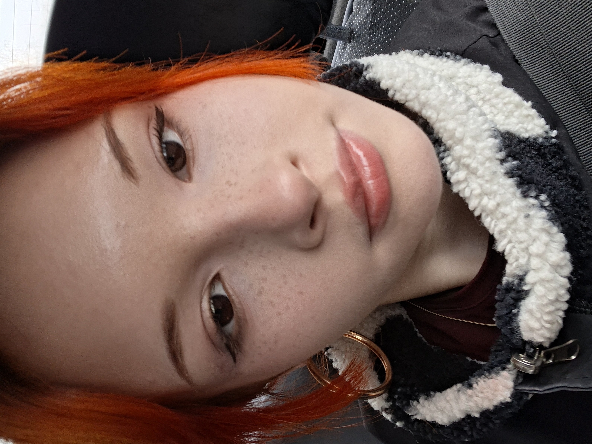 The image is a close-up of a woman with bright orange hair. She has light skin, brown eyes, and freckles on her face. She is wearing eyeliner and lip gloss. Her face is partially obscured by the collar of a black and white fleece jacket. A gold hoop earring is visible. A thin gold necklace can be seen under the jacket. There is no information to determine that this image was taken in the city of Chaikovsky, Russia.