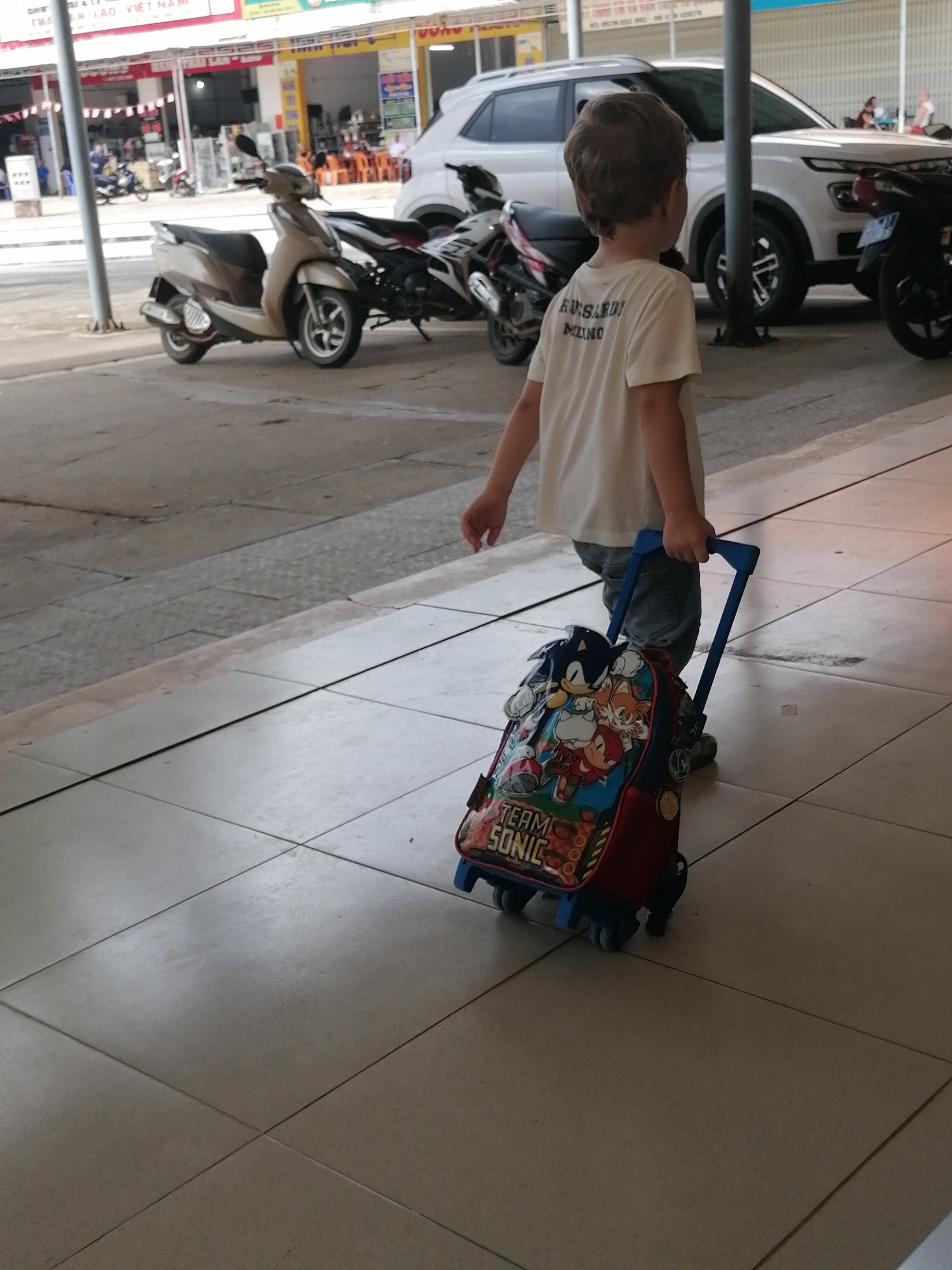 A young boy with brown hair is walking away from the camera, pulling a rolling backpack with a Sonic the Hedgehog design. He is wearing a white t-shirt with text on the back and blue jeans. The backpack is red and blue with images of Sonic, Tails, and Knuckles. The boy is walking on a tiled sidewalk in front of a building. In the background, there are several parked motorcycles and a white SUV. The scene appears to be in a street or commercial area in Bo Y, Vietnam, as indicated by signage in Vietnamese on the buildings. The lighting suggests it is daytime, likely sunny.