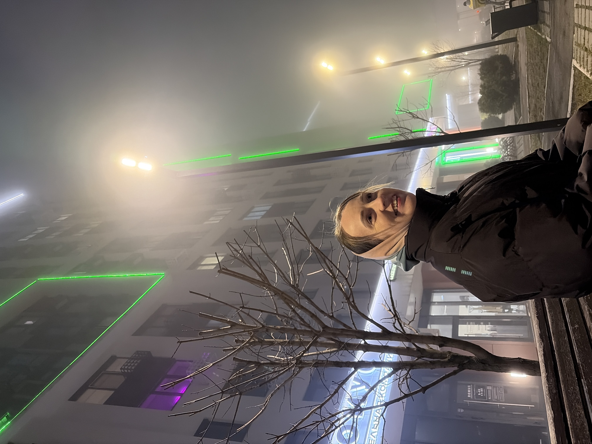 A female is sitting on a park bench in Izhevsk, Russia on a foggy night. A building is in the background with green and purple neon lights, and street lights. There is also a bare tree next to the bench.