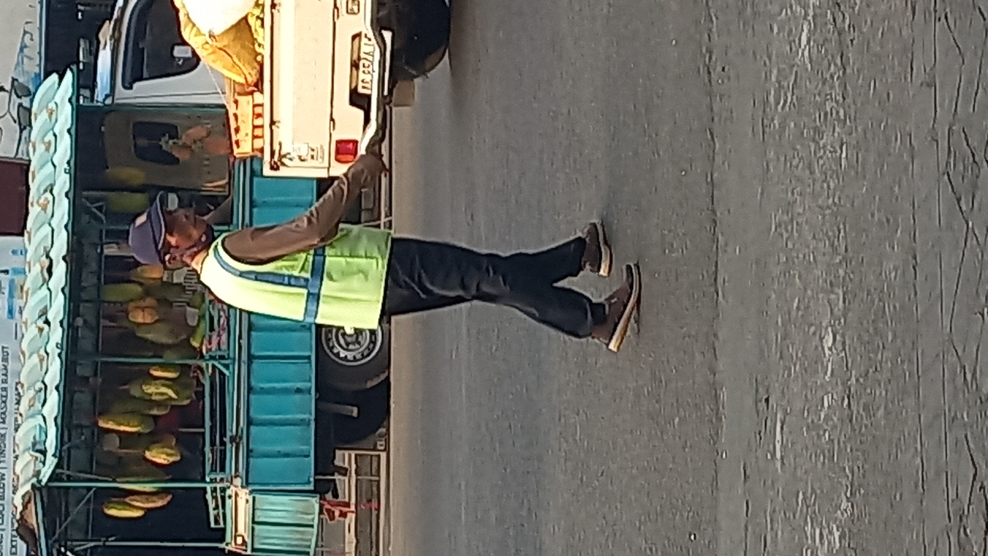 A person wearing a dark blue cap, a light brown long-sleeved shirt, a bright green safety vest with blue reflective stripes, and dark pants walks on an asphalt road. The person is also wearing dark brown athletic shoes with orange accents. Behind the person is a turquoise truck with a green canopy, laden with what appear to be durian fruits. To the right of the truck is a white truck with a yellow sack in the bed and a license plate visible that reads "IC 9526 IA". The scene appears to be an outdoor market or roadside vendor location in Mangunsari, Indonesia. The lighting suggests it is daytime.