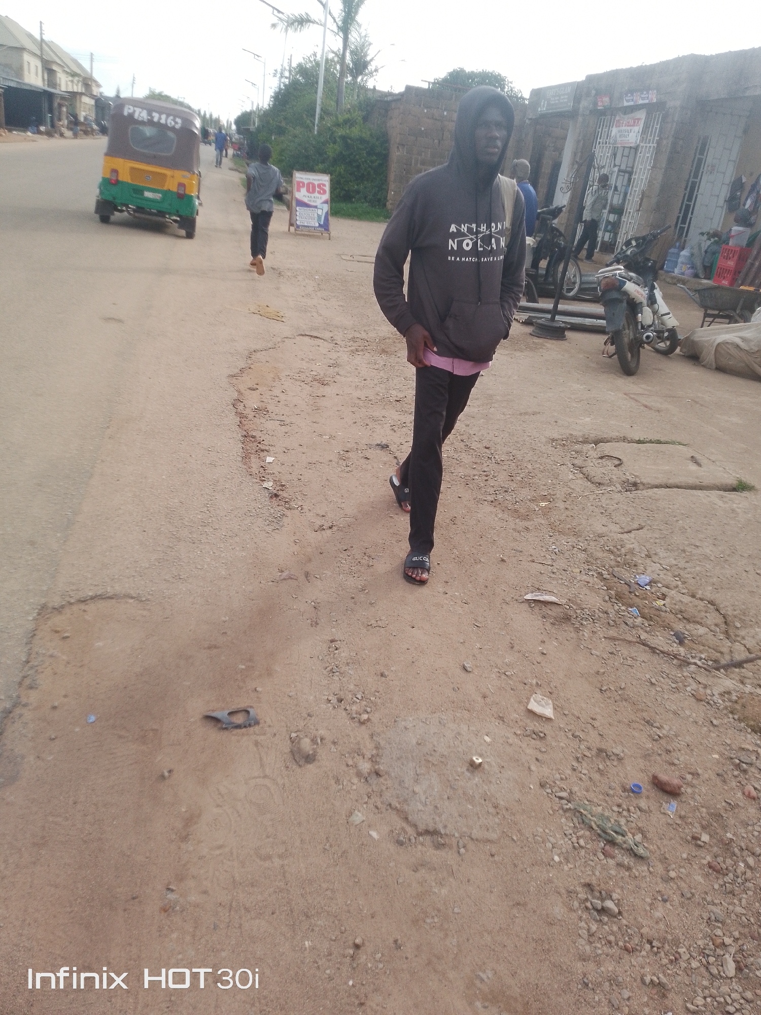 The image depicts a street scene in an urban environment, possibly in a developing country. A young man, wearing a dark hoodie with white text and pink undershirt, black pants, and black slides, is walking towards the camera. He has a beige strap across his shoulder. In the background, a yellow and green auto-rickshaw is visible, with "PTA-2163" on its rear. To the right of the auto-rickshaw, a sign with "POS" and other text is placed on the side of the road. Several motorcycles, one red and white, are parked in front of buildings. The ground is dusty and uneven, with scattered debris. The buildings appear to be made of brick and have corrugated metal roofs. The sky is overcast. The overall impression is one of a busy, informal commercial area.