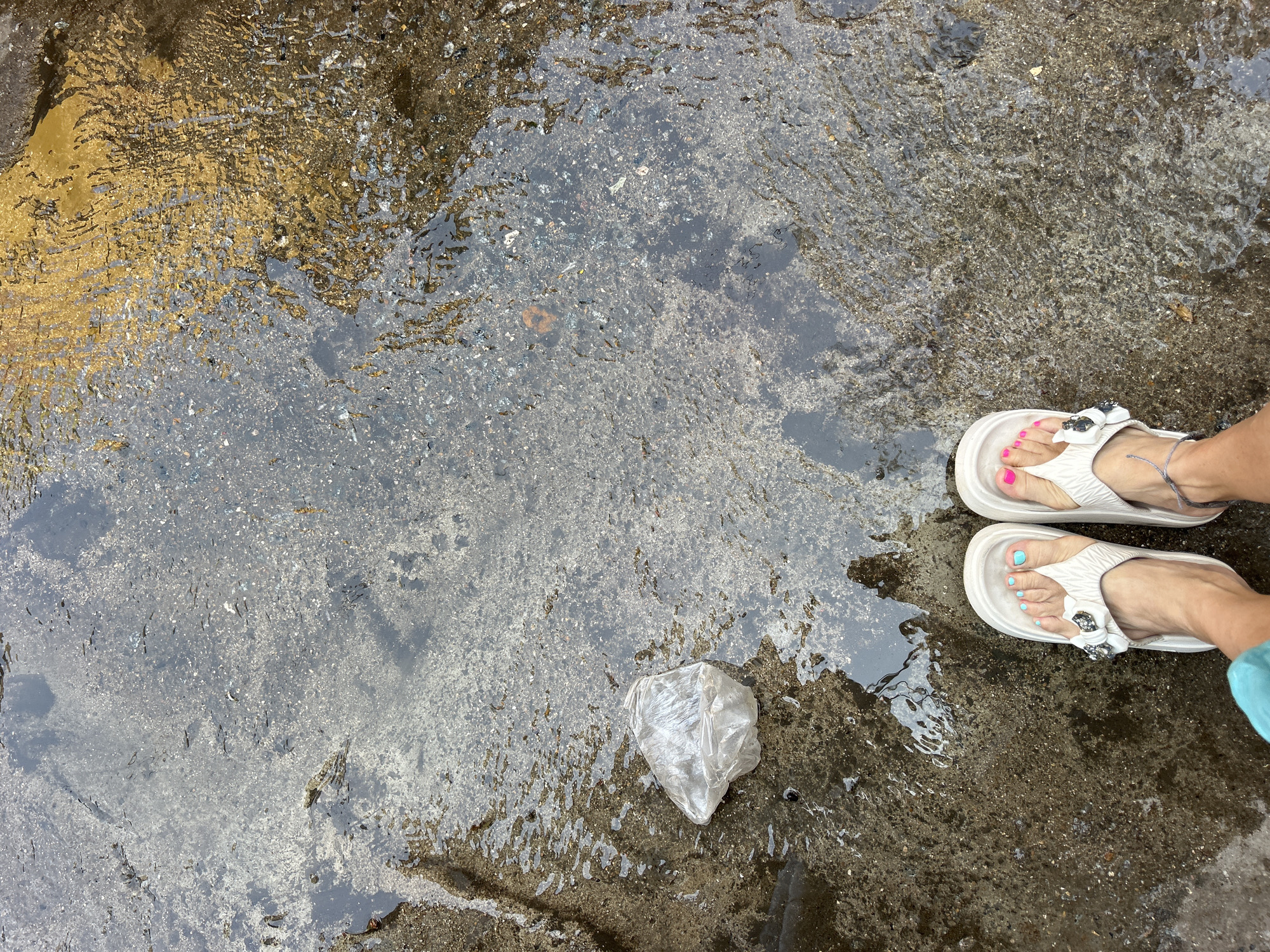 A high-angle, full shot depicts a waterlogged concrete surface in Puttaparthi, India. The left third of the frame shows a yellow-tinged section. The remaining area is covered in a reflective layer of water, beneath which the rough texture of the concrete is visible. A crumpled, clear plastic bag sits in the water near the center of the frame. In the upper right corner, a pair of feet wearing white sandals are positioned. The toenails are painted in contrasting colors of pink and turquoise. The sandals have decorative floral embellishments. A small portion of turquoise-colored clothing is visible on the right edge.