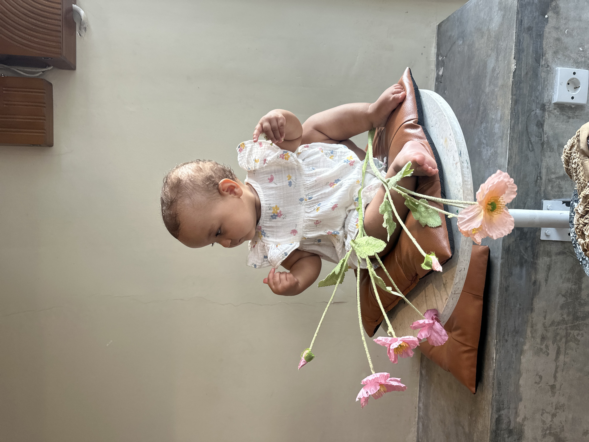 A baby sits on a cushion and a white wooden object in North Kuta, Indonesia. The baby is wearing a white, floral-patterned dress and is holding a bunch of light pink flowers. The seat is constructed of a brown cushion on a white, round platform. The setting appears to be indoors, with a light beige wall to the left and a gray cement wall to the right. There is an electrical outlet on the cement wall.