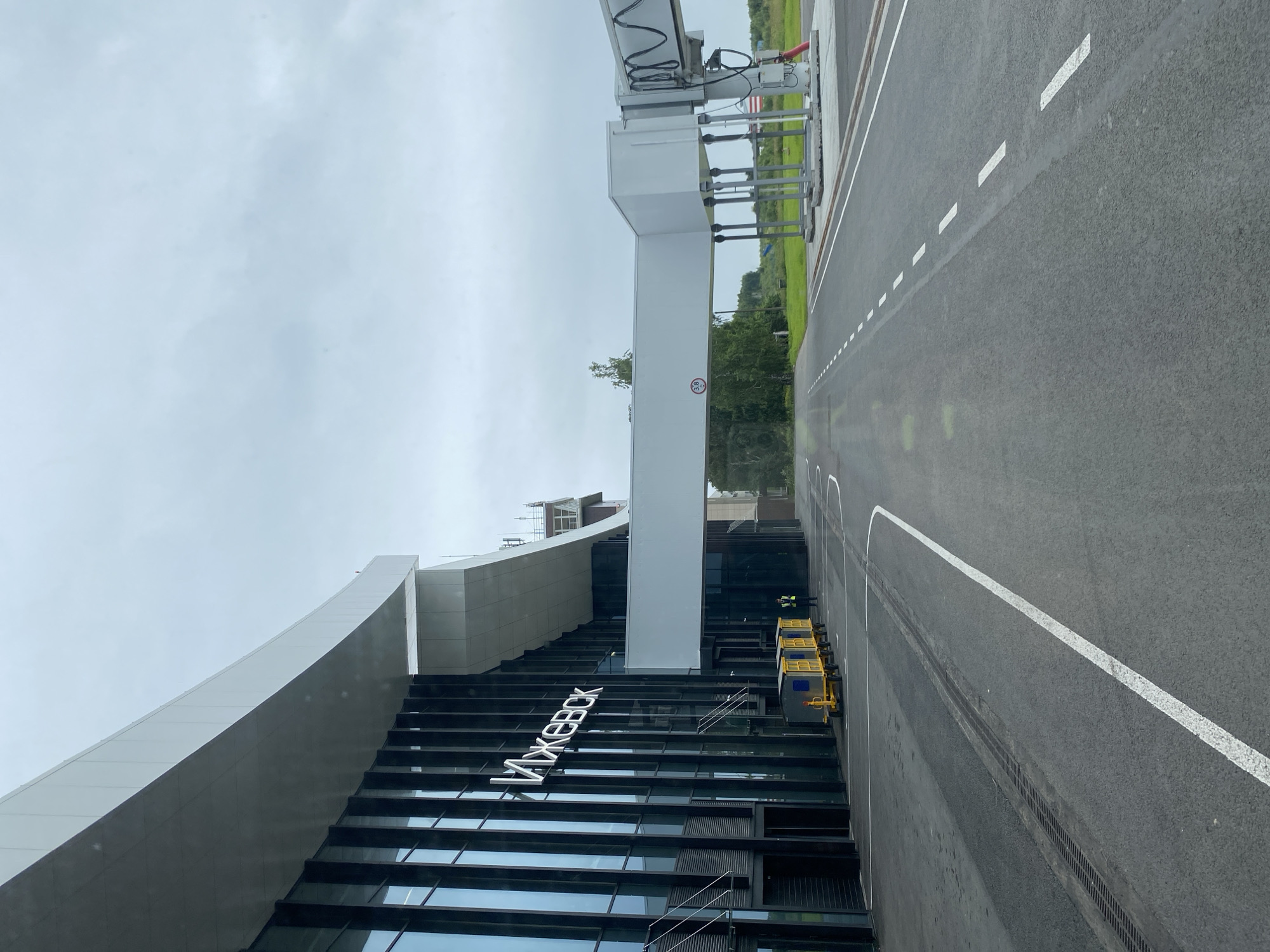 The image shows the exterior of an airport terminal building with "ИЖЕВСК" written in large white letters on the facade. A jet bridge extends from the terminal to an unseen aircraft or gate. In front of the terminal, several yellow baggage carts are lined up, and a person wearing a high-visibility vest stands nearby. The ground in front of the building is asphalt with white lane markings. The sky is overcast, and trees are visible in the background, suggesting a rural or semi-rural location. The specific location is the airport in Novoye Martyanovo, Russia.