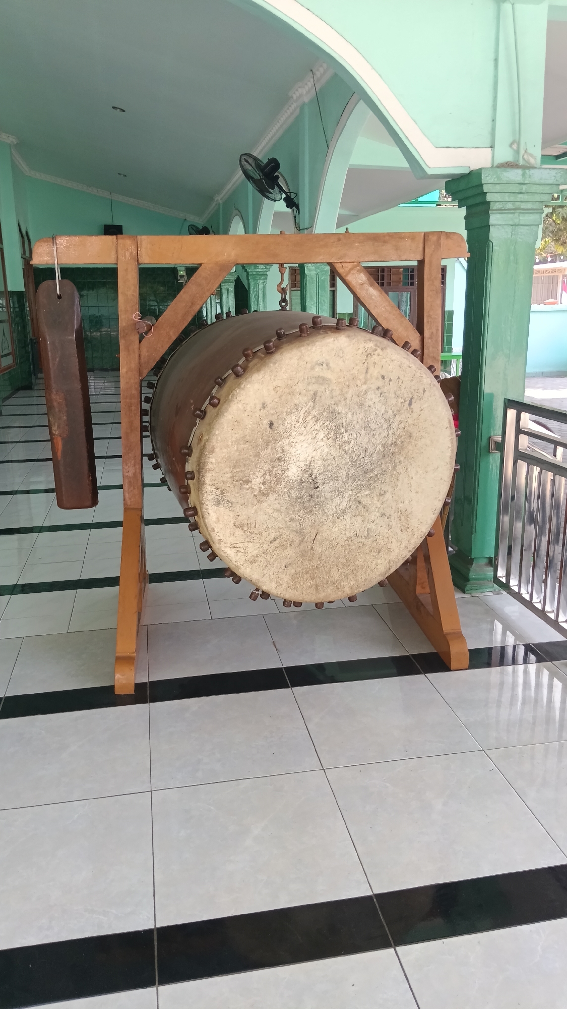 A large, round drum with a pale, textured surface is centrally positioned in the image, resting on a wooden stand. The drum is adorned with numerous small, dark, circular fasteners around its circumference. To the left of the drum, a long, dark, wooden object hangs from a rope, also suspended from the wooden stand. The scene appears to be an outdoor covered area, possibly a veranda or portico, indicated by the tiled floor with alternating light and dark bands. In the background, there are green-painted pillars and a metal railing. A black oscillating fan is mounted on the wall above and behind the drum. The overall color palette is muted, with dominant tones of beige, brown, and green. The lighting suggests a daytime setting. This image was captured in Purwokerto, Indonesia.