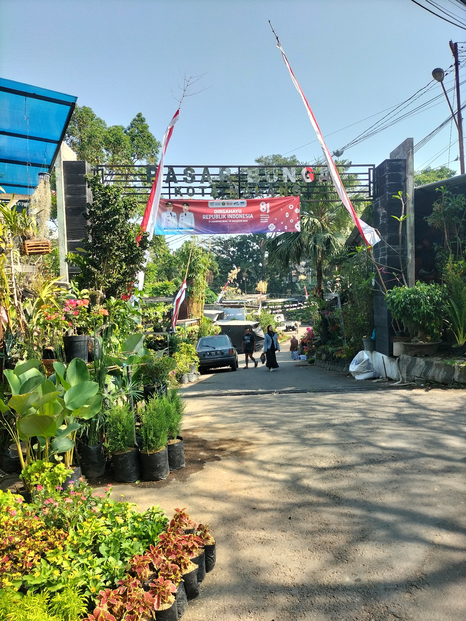The image depicts the entrance to "PASAR BUNGA KOTA" in Malang, Indonesia. A red banner with "DIRGAHAYU REPUBLIK INDONESIA" and images of two men in uniform hangs above the entrance, suggesting a celebration of Indonesia's Independence Day. Red and white Indonesian flags are prominently displayed, stretched diagonally across the entrance. The area is a bustling flower market, with numerous plants and flowers arranged for sale in black plastic bags and pots. A grey car is parked on the road, and two individuals are walking away from it. The ground is a mix of asphalt and dirt, with shadows from trees cast across it, indicating an outdoor setting in daylight. The background reveals lush greenery and other market stalls.