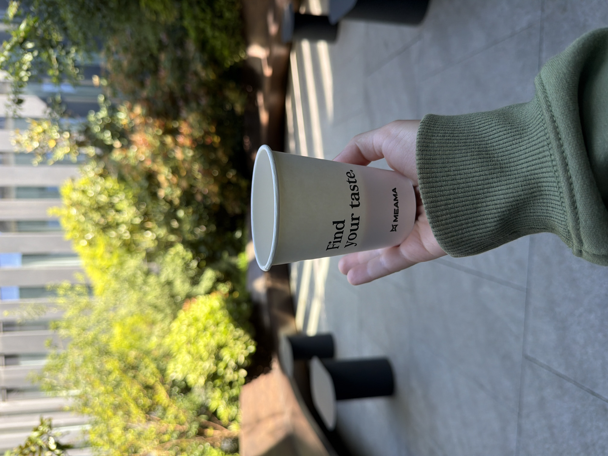 A vertical-oriented, close-up shot captures a person's hand holding a white paper cup outdoors. The hand, extended from the right side of the frame, is wearing a long-sleeved garment with a ribbed cuff in a muted olive green color. The paper cup is the primary object in focus, displaying black text that reads "Find your taste." vertically aligned on its side. Below this phrase, a logo featuring a star-like symbol precedes the word "MEAMA". The cup appears empty.
The background is significantly blurred, creating a soft bokeh effect. It features abundant green foliage from trees or bushes, suggesting a natural or landscaped urban environment. Behind the greenery on the left, a modern building with light-colored, horizontally structured elements is faintly visible. The ground beneath the hand is a light-colored paved surface, with a few dark, cylindrical objects, possibly bollards or decorative elements, visible further in the blurred background. The bright natural lighting indicates it is daytime and sunny. The scene depicts a casual moment in an outdoor urban setting, potentially a park or a commercial area in Tbilisi, Georgia. No specific activity beyond holding the cup is evident, and no emotions are discernible.