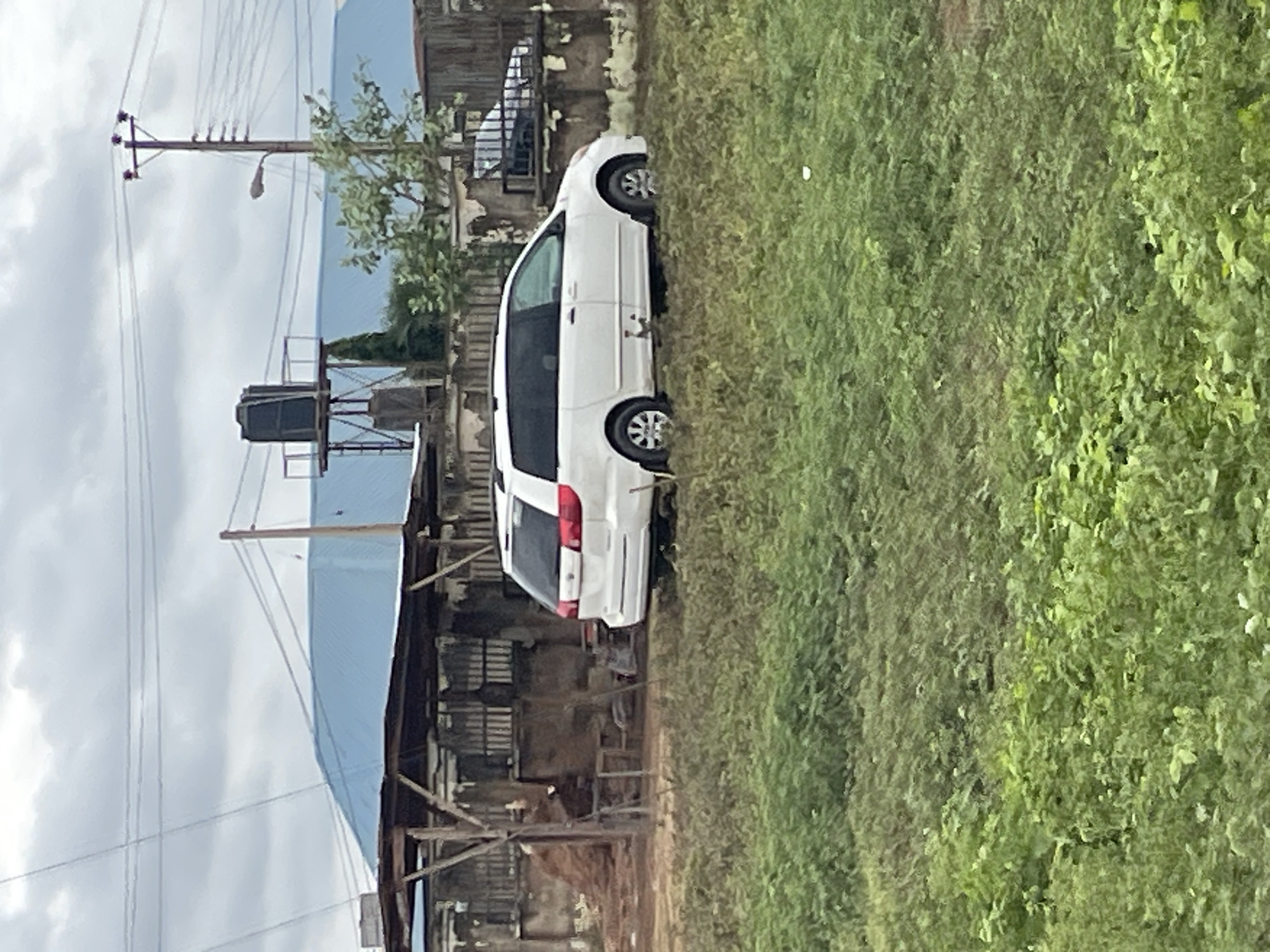 A white minivan is parked on a grassy area in front of a makeshift structure with a corrugated metal roof supported by wooden posts. Behind the structure, a building with a blue, sloped roof is visible, along with a large black water tank on a metal platform and a utility pole with multiple wires. A second, darker vehicle is partially visible behind a low wall on the right side of the image. The foreground is dominated by dense green vegetation. The sky is overcast. The scene is located in an outdoor, semi-urban or rural setting, possibly in Nigeria based on the water tank and building style.