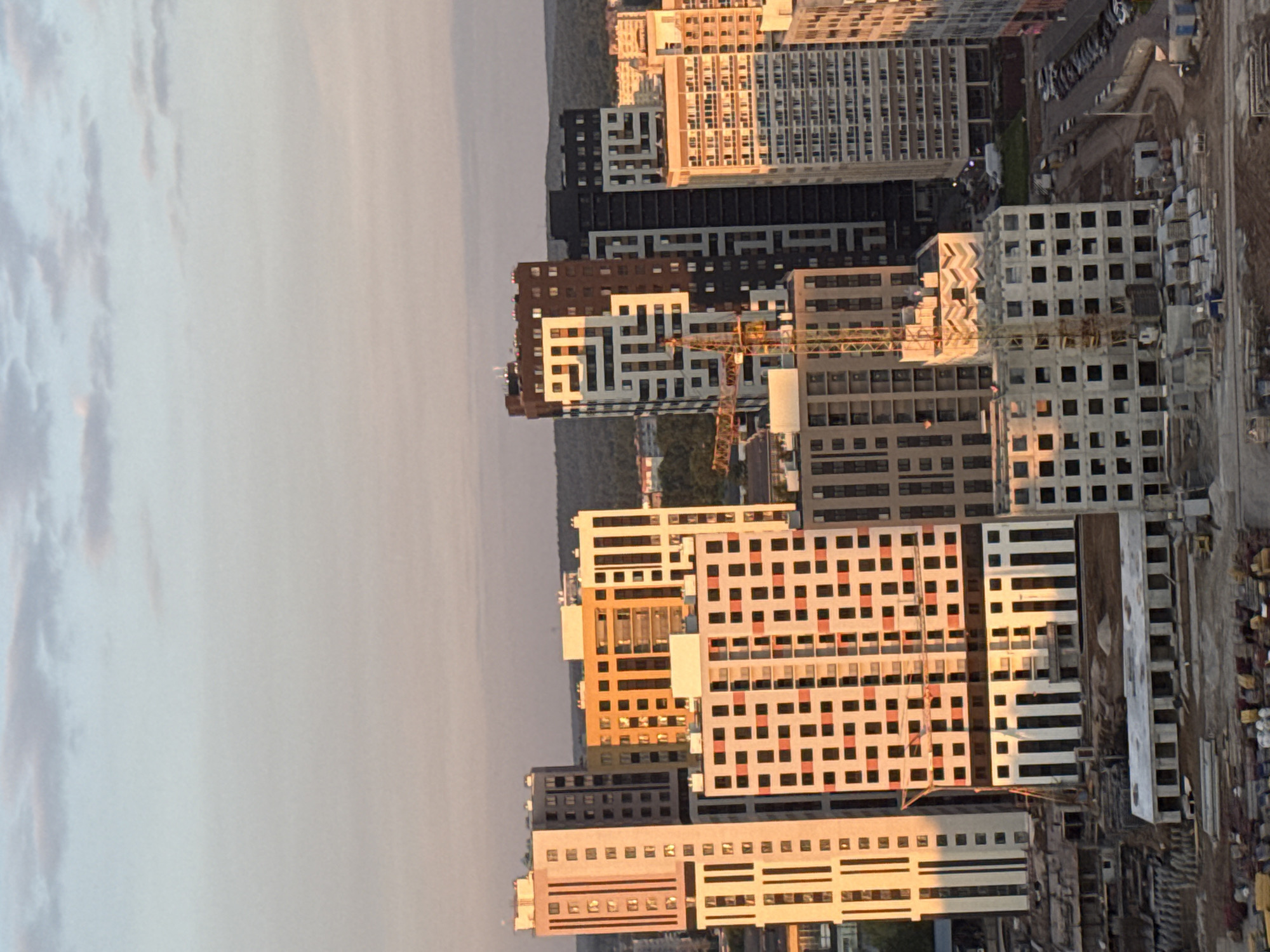 The image captures a modern urban landscape at either sunrise or sunset, indicated by the warm, golden light casting long shadows across numerous multi-story buildings. The scene primarily features residential structures, many exhibiting contemporary architectural styles with varying facade colors including white, black, brown, orange, and panels with red accents.
Several buildings appear to be under active construction or recently completed, with at least one prominent yellow construction crane visible among the structures. Piles of raw building materials are scattered on the ground near these sites. A paved road or parking lot with several cars can be seen in the mid-ground, alongside further construction activity.
In the background, a dark, undulating landscape, possibly consisting of hills or dense tree cover, stretches towards the horizon under a soft, cloudy sky. The overall setting portrays a burgeoning urban development, consistent with new residential areas. Given the provided context, the location is Perm, Russia. There is no visible, legible text in the image.