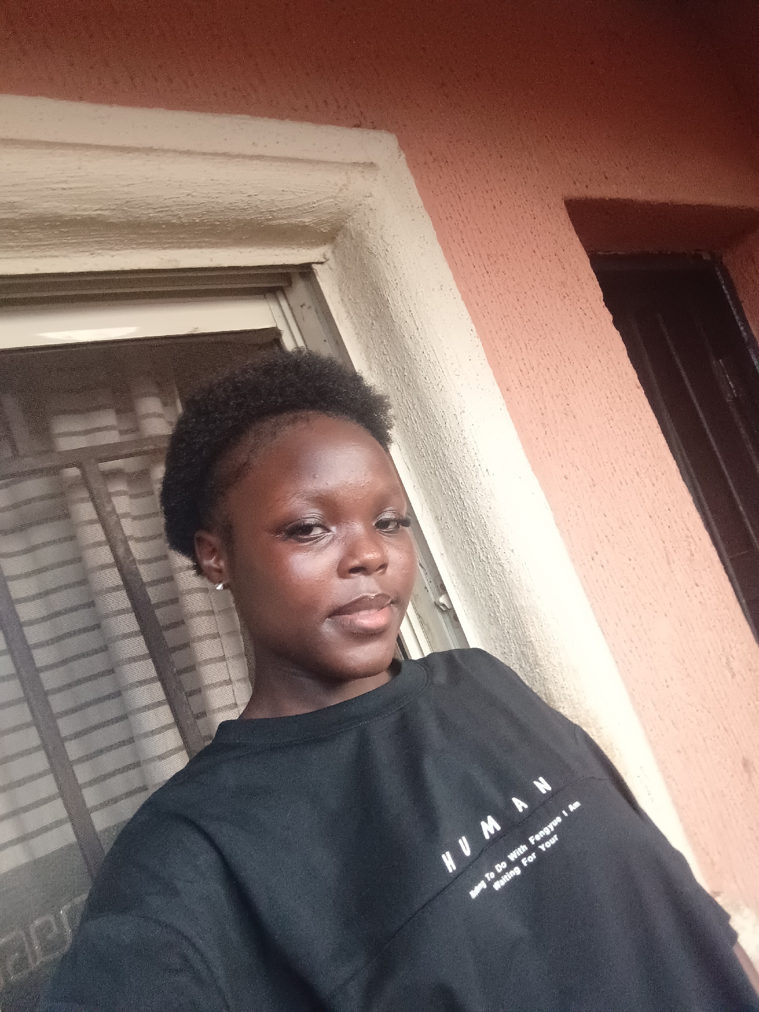 A young woman with dark skin and short, dark hair poses for a selfie in Onitsha, Nigeria. She wears a black t-shirt with the word "HUMAN" printed on it. She is standing next to a window with metal bars and a peach-colored wall. The light suggests it is daytime.