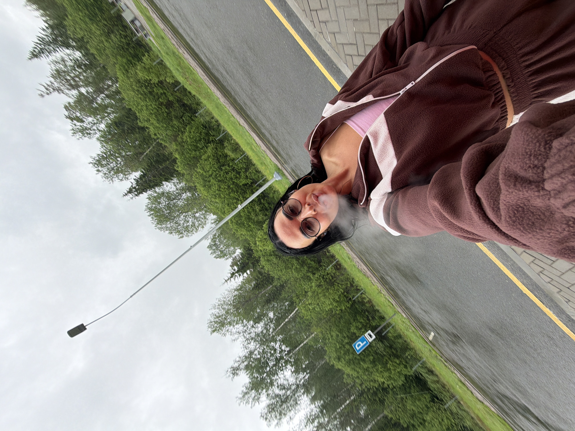The image shows a person standing beside a street in Sust'e-Konets, Russia. The individual, wearing sunglasses and a brown fleece jacket over a pink top, is exhaling vapor, likely from an e-cigarette or similar device. Behind the person is a road with a yellow line marking, followed by a grassy area and trees. A street light and a parking sign are also visible. The weather appears overcast.