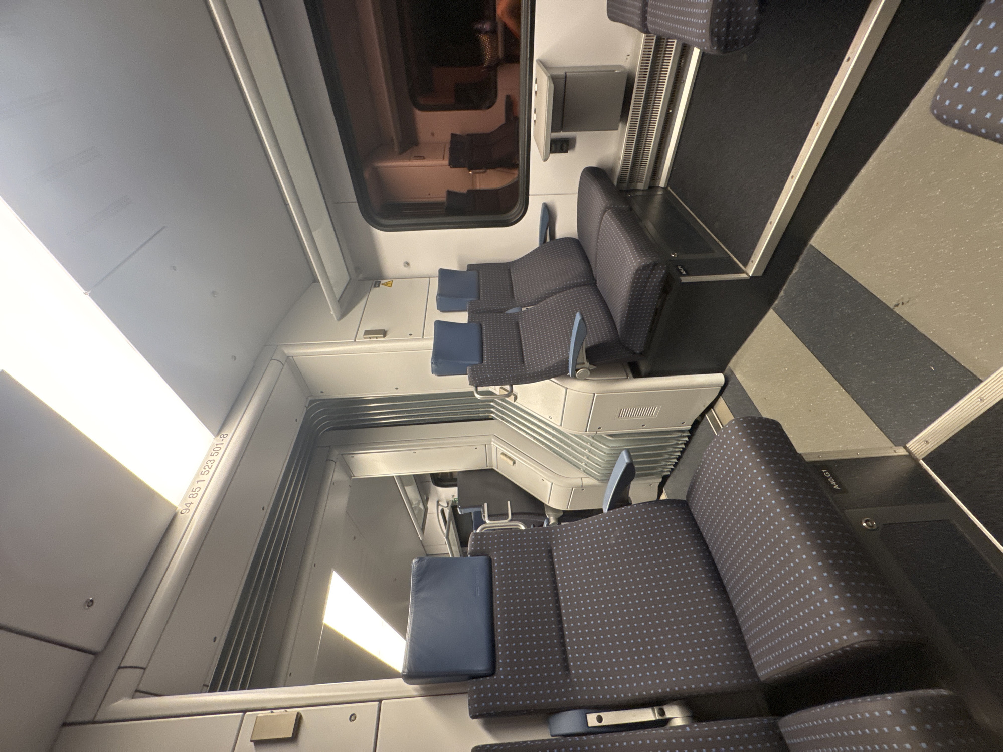 The image shows the interior of a train car. The seats are upholstered in a dark fabric with a small blue dot pattern. There are headrests on the seats. The floor is a speckled gray and black material. There is a window on the right side of the image, showing reflections of lights and possibly other train cars. A small tray table is attached to the wall near the window. In the background, there are more seats and a doorway leading to another section of the train. The lighting appears to be artificial, coming from overhead fixtures. There are no people visible in the image. The overall environment is clean and modern. The image was taken in Val Mara, Switzerland.