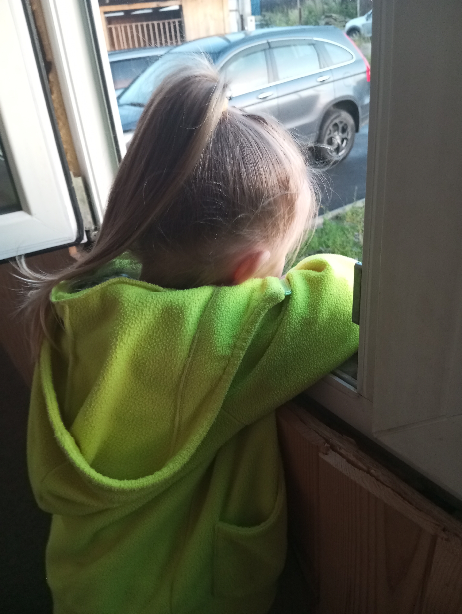 A young child, seen from behind, is looking out of a window. The child has blonde hair pulled into a ponytail and is wearing a bright green hooded jacket. The window frame is white, and beyond the window, a silver SUV is parked. The surrounding environment suggests a residential or suburban area.