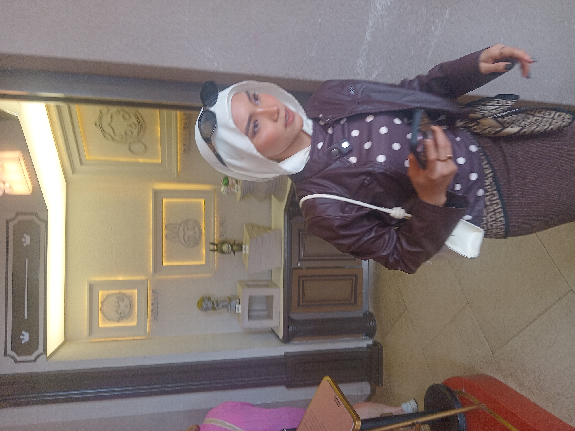 The image captures a woman wearing a white hijab, sunglasses perched on top, a maroon polka-dot shirt, and a matching maroon jacket. She is holding a smartphone in her right hand and has a white purse slung over her left shoulder. The background features decorative frames on a wall, some with figures inside, and display shelves with figurines and books. The setting appears to be an indoor retail or exhibition space, possibly in Da Nang, Vietnam, given the context. The flooring is tiled.