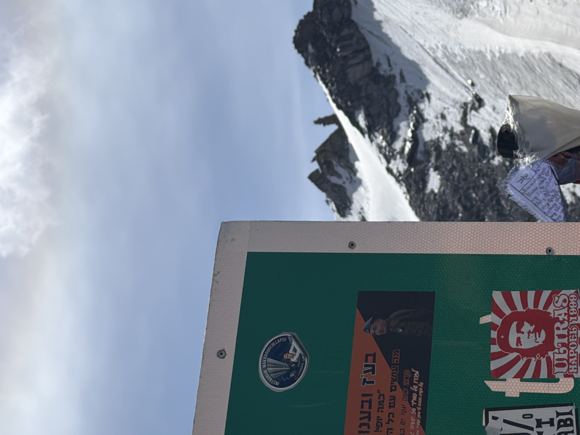 The image shows a close-up view of a green sign mounted on a light-colored surface, possibly a wall or a structure, with a mountainous, snow-covered landscape in the background. The sign has several stickers and text in Hebrew. One circular sticker at the top features a depiction of a person with the text "INSTAGRAM MAXXOOOCOLLAPSE" and "LADAKH, INDIA". Below this, there's a larger sticker with text and an image of a person in military attire. Further down, a red and white sticker shows a portrait of Che Guevara with "ULTRAS HAPOEL 1999" printed below. In the background, rugged mountain peaks are visible, partly covered in snow, under a bright sky with clouds. The overall scene suggests a location in a mountainous region, with the presence of specific cultural and political references on the sign.