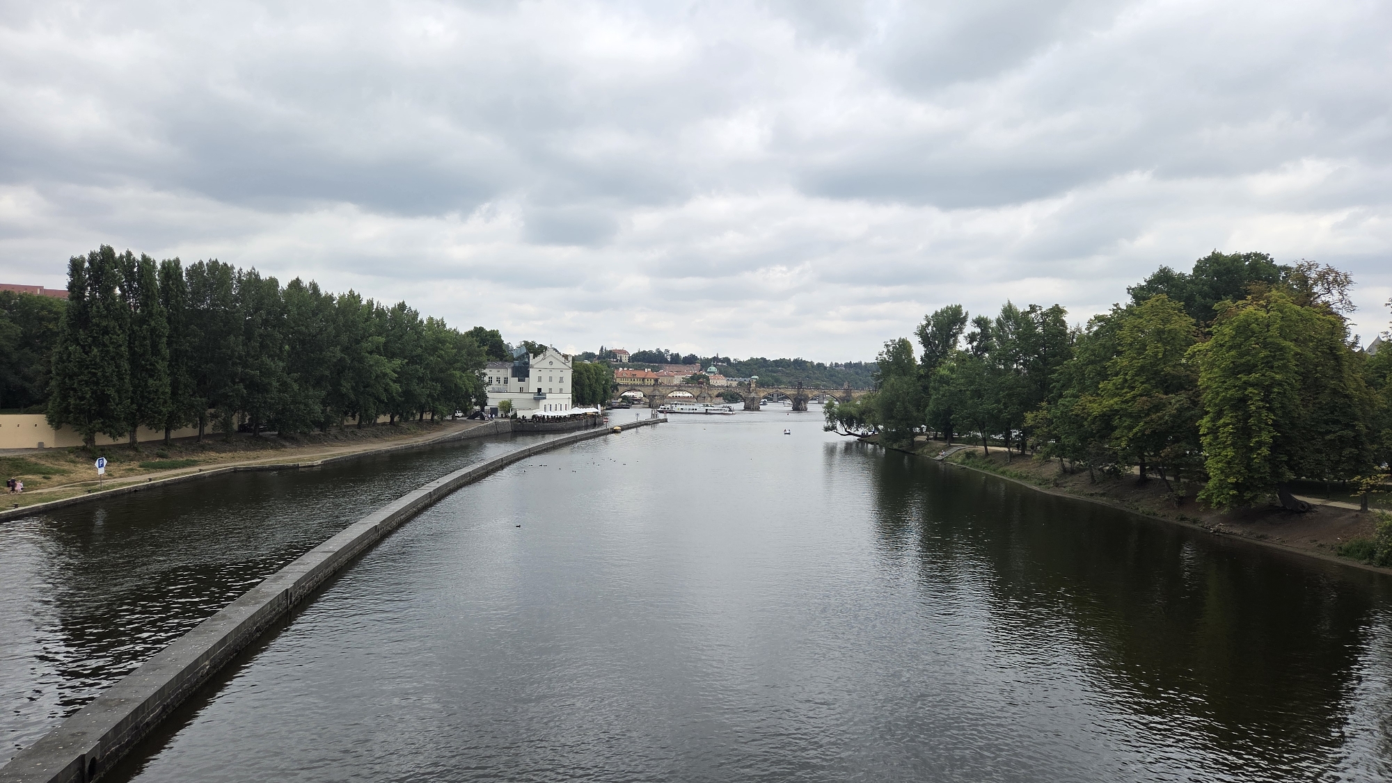 The image shows a wide river flowing through the city of Prague, Czechia. The river is calm and reflects the cloudy sky above. On the left bank of the river, there is a concrete embankment, followed by a grassy area with trees and a path. A white building with a red roof is visible on the bank. Further in the distance, a long stone bridge with multiple arches spans the river, and beyond that, more buildings and a hill with a castle-like structure are visible. On the right bank, there are numerous trees and a path running alongside the river. There are a few small boats on the water and some people are visible on the left bank. The overall scene depicts a tranquil, urban landscape.