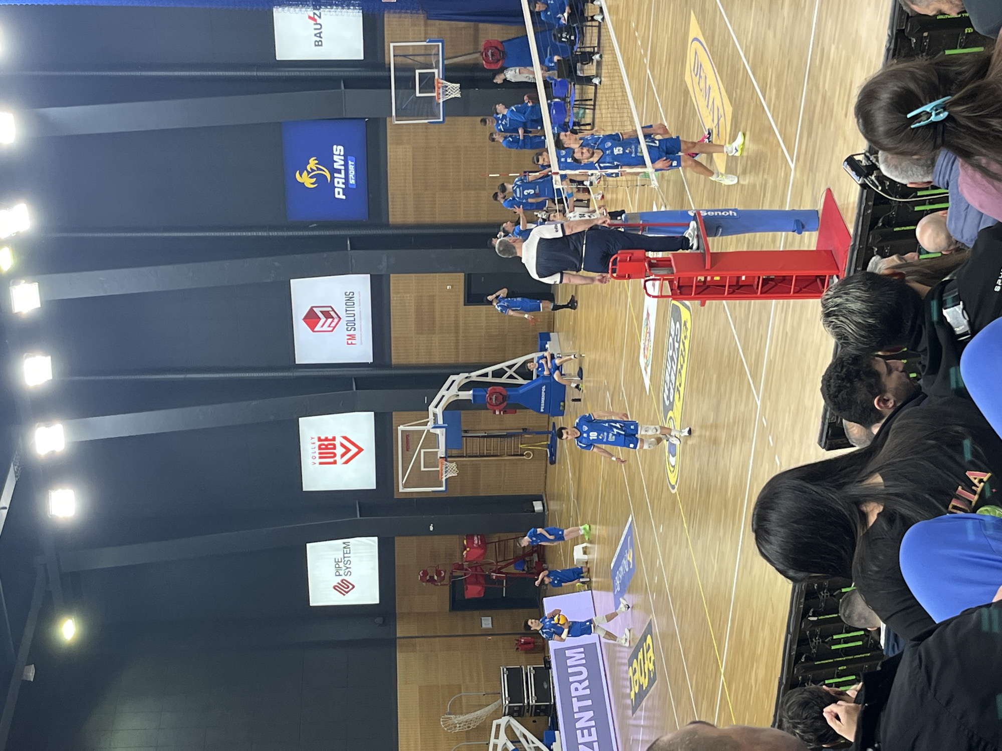 Here's a forensic description of the image:
**Scene Overview:**
The image appears to be taken from the stands at a volleyball game. The court is well-lit. A volleyball net divides the court in the center. There are players on both sides of the net, dressed in blue uniforms.
**People:**
* There are multiple volleyball players visible on the court, predominantly wearing blue uniforms. Some players appear to be warming up or practicing.
* There are spectators in the foreground, including a woman with a hair clip. Several other spectators can be seen in the background on the sidelines.
* A coach in a white/black collared shirt is standing on the sidelines.
**Objects:**
* Volleyball net.
* Referee stand (red).
* Volleyballs.
* Banners with logos/brands hanging from the ceiling: "BAUZIT", "PALMS SPORT", "FM SOLUTIONS", "VOLLEY LUBE", and "PIPE SYSTEM".
* Floor advertisements for "ZENTRUM" and "Ebet."
**Actions/Interactions:**
* Volleyball players are engaged in practice activities.
* Spectators are watching the game.
**Environment:**
* The location is indoors, presumably a sports arena or gymnasium.
* The court is made of wood.
* Based on the sponsor advertisement banners, this sporting event is taking place in Sofia, Bulgaria.