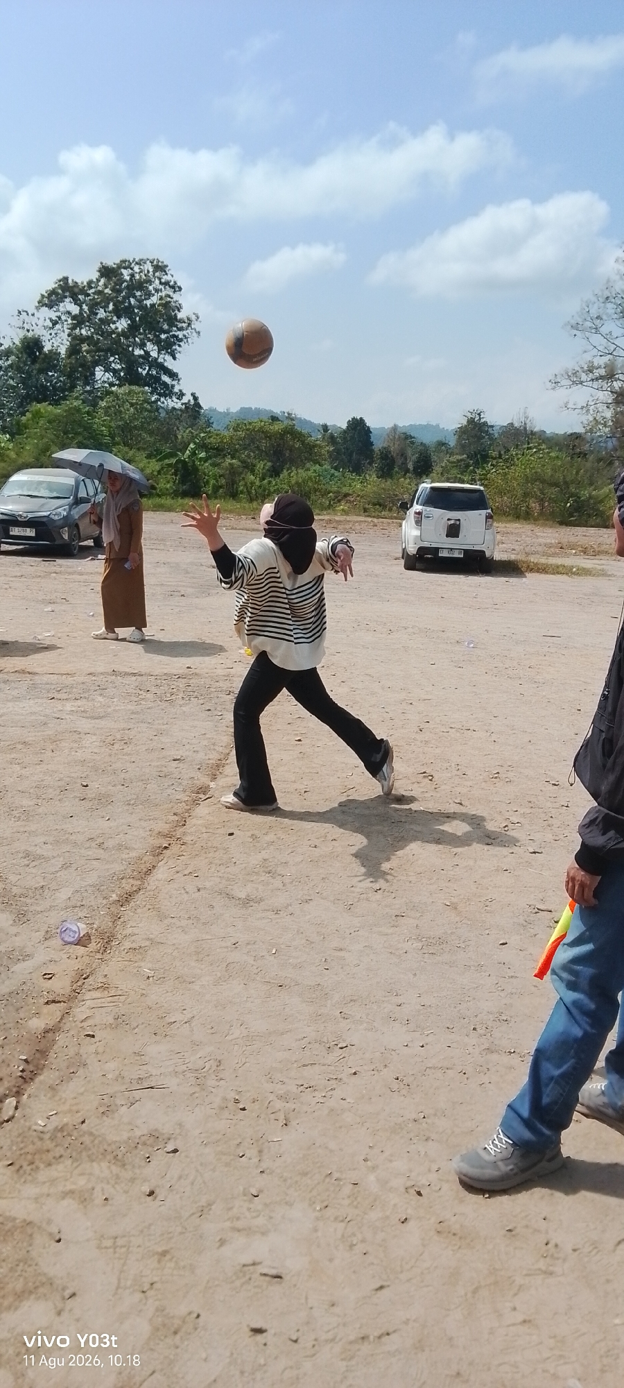 The image depicts an outdoor scene, possibly in Tirawuta, Indonesia, on a sunny day with a partly cloudy sky. In the foreground, a person wearing a striped long-sleeved shirt, black pants, and a headscarf is in mid-action, appearing to be hitting or tossing a volleyball. Their left arm is extended upwards, and their right arm is bent, suggesting they are in the process of serving or spiking. The volleyball is suspended in the air above them. To the left, another person, also wearing a headscarf and a light brown dress, is standing under a large umbrella and observing the activity. In the background, several vehicles are parked, including a dark-colored car on the left and a white SUV on the right. The ground is dry and dusty, with some scattered debris. The environment is characterized by trees and vegetation in the distance, with rolling hills forming the horizon. A portion of another person's legs, wearing blue jeans and grey sneakers, is visible on the right side of the frame, and they are holding a colorful object, possibly a toy or a marker.