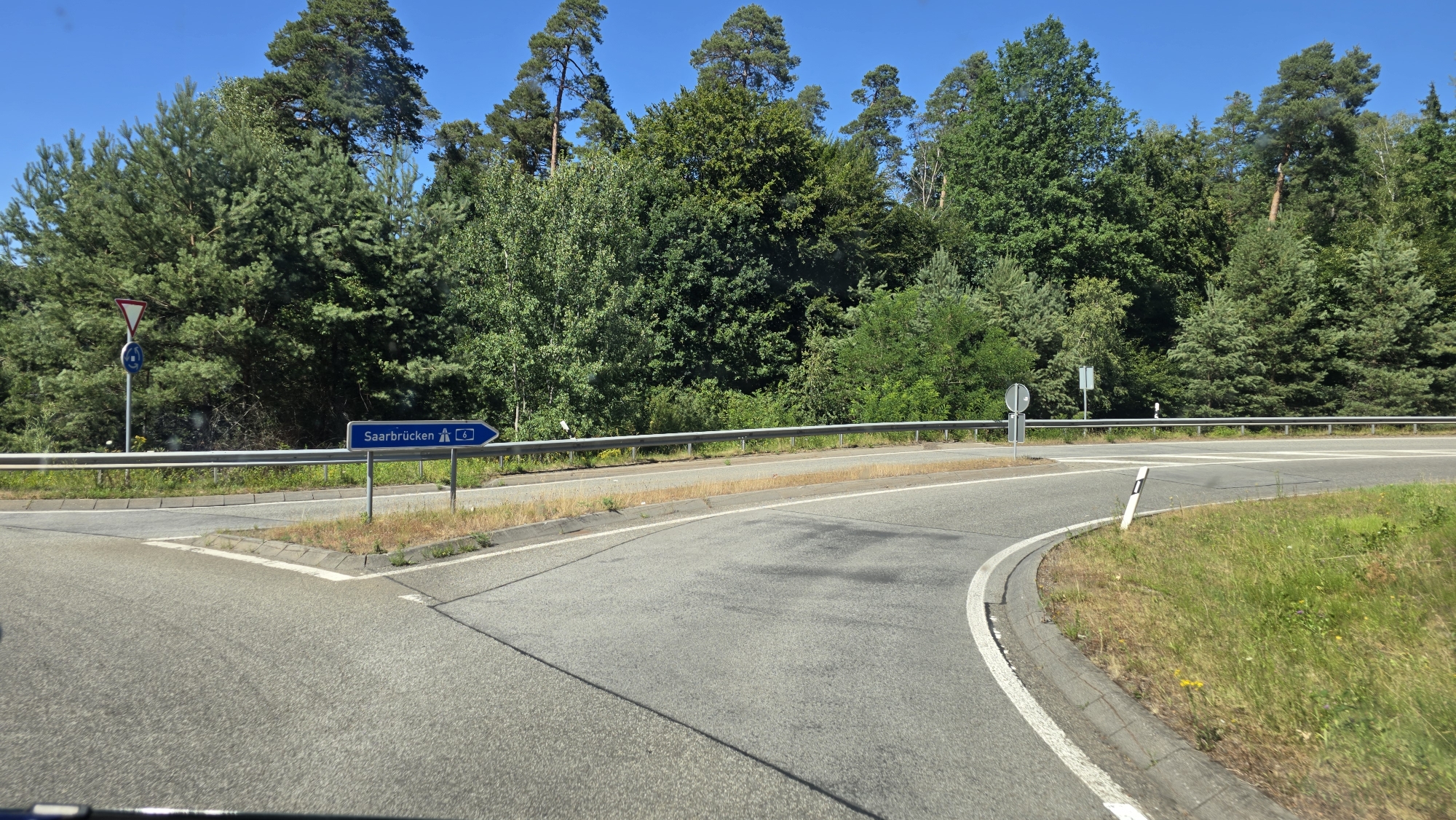 The image shows a road junction on a clear, sunny day with a bright blue sky. The road surface is asphalt, with white lines marking the lanes. On the left side of the road, there is a blue sign with white text and a symbol indicating a direction towards "Saarbrücken" and the number "6". To the left of this sign, there is a white triangular warning sign with a red border and a black symbol inside, followed by a circular blue sign with a white symbol. A metal guardrail runs along the side of the road. The background is dominated by dense, green trees, with a mix of coniferous and deciduous species. There are no people visible in the image. The scene appears to be a highway exit or a junction on the outskirts of a wooded area. This is a description of the junction of the B40 and the L502 near Kaiserslautern, Germany.