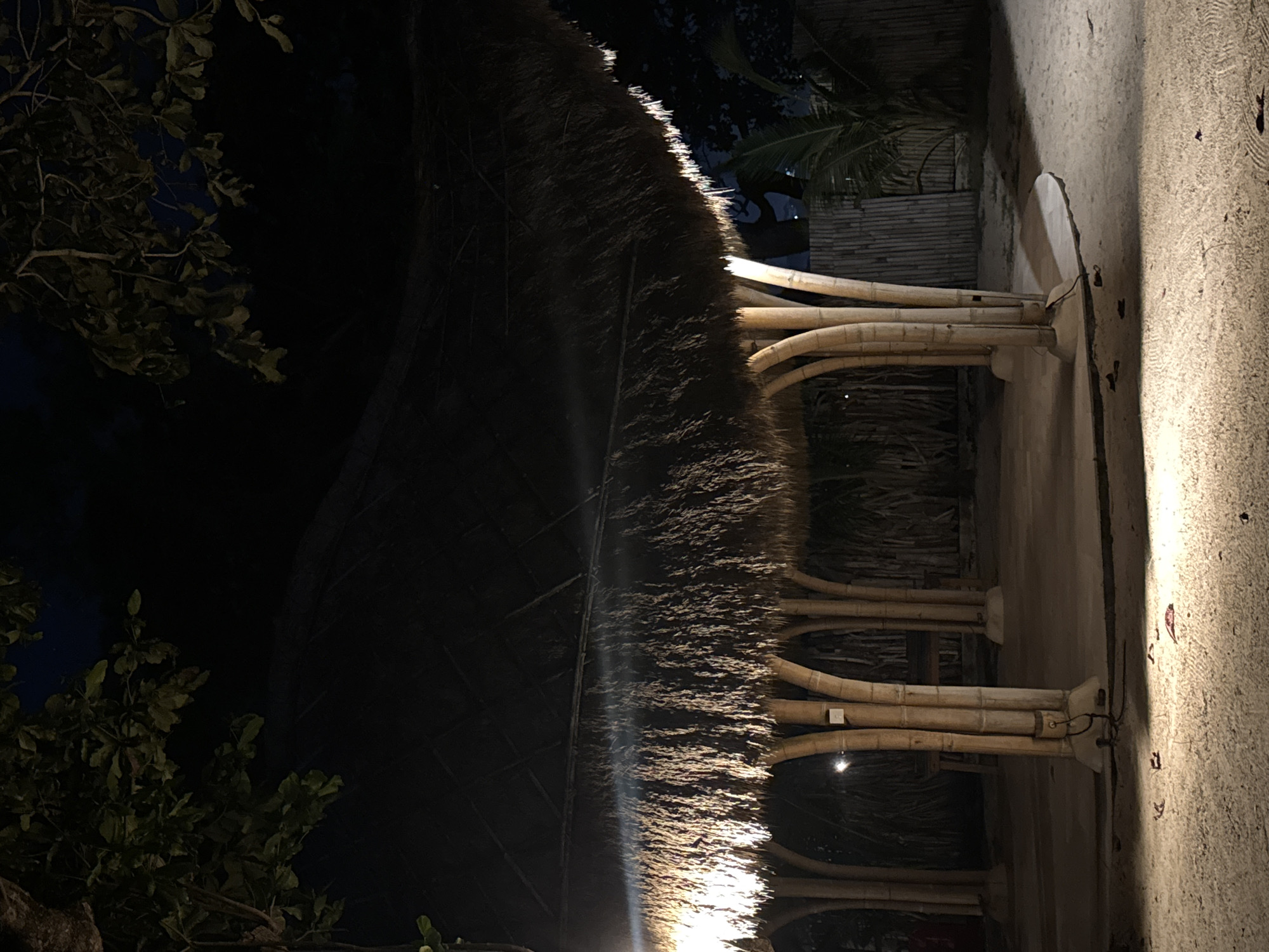 The image, taken at night in Kuta Selatan, Indonesia, depicts an outdoor tropical structure. No people are visible.
The central feature is a thatched roof, dramatically illuminated from below by artificial lights, causing bright highlights to streak across its shaggy texture. The roof curves gracefully, creating a sense of an open-air pavilion or sheltered walkway. Supporting this roof are several clusters of sturdy bamboo columns, each comprising multiple individual bamboo stalks joined together. These columns exhibit a smooth, light brown texture and are also highlighted by the upward-facing lights.
To the right, a textured concrete or stucco surface, likely a ground or a low wall, is brightly lit, revealing a coarse, somewhat uneven finish. Some small debris, possibly leaves or dirt, is scattered on this surface. Electrical wiring is visible connecting small light fixtures at the base of the bamboo supports.
In the background and to the left, dark tropical foliage, including leaves and branches from trees and bushes, is silhouetted against a dark night sky, hinting at the lush environment. The overall scene is characterized by strong contrasts between deep shadows and bright, warm artificial lighting, creating an inviting and serene nighttime ambiance typical of a resort or restaurant setting. No visible text is present in the image.