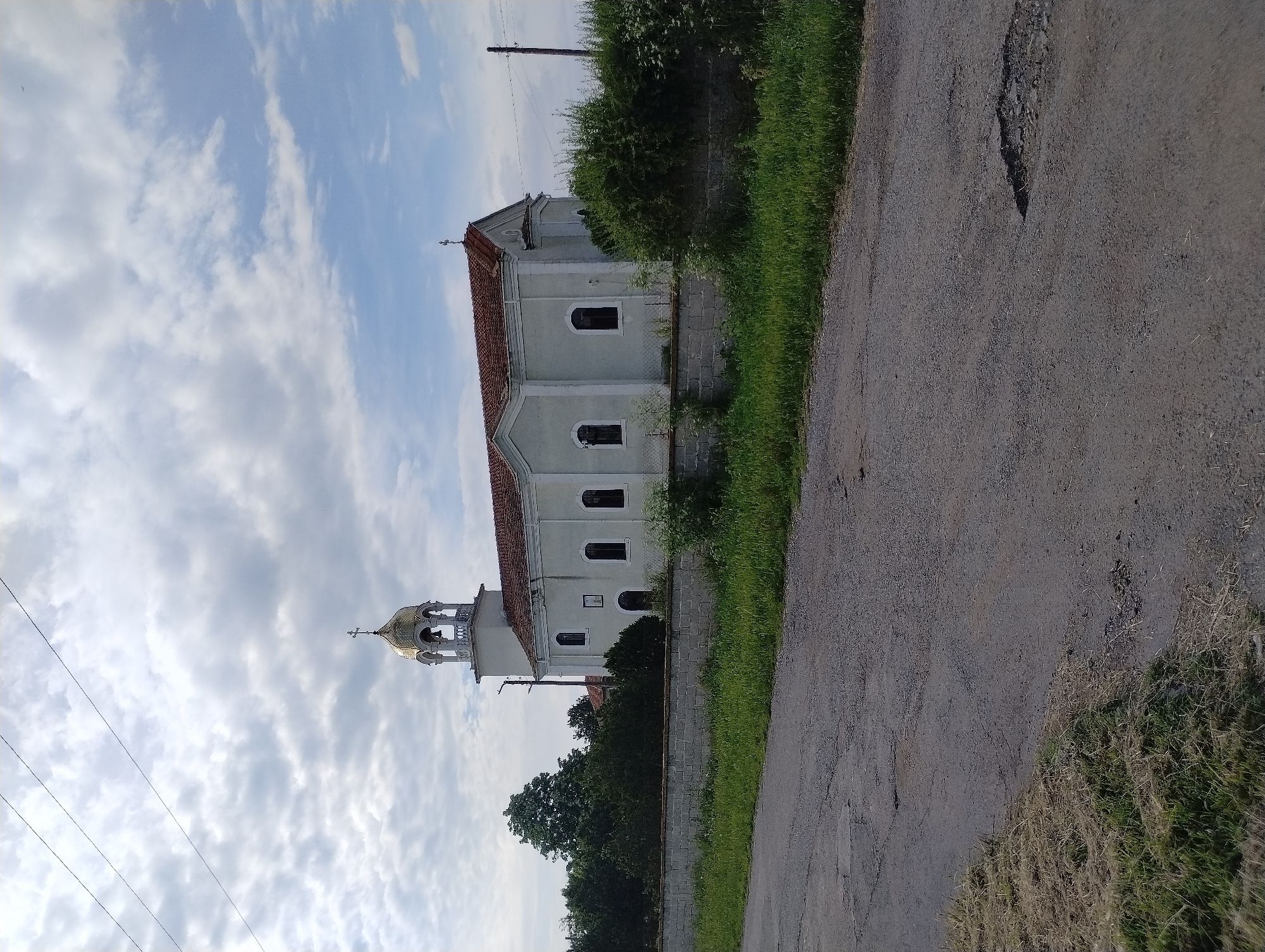 The image shows a church in Roza, Bulgaria. The church is light-colored with a red tile roof and a gold-domed bell tower. It stands next to a paved road, with vegetation along the roadside. The sky is cloudy. Power lines are visible above the church.