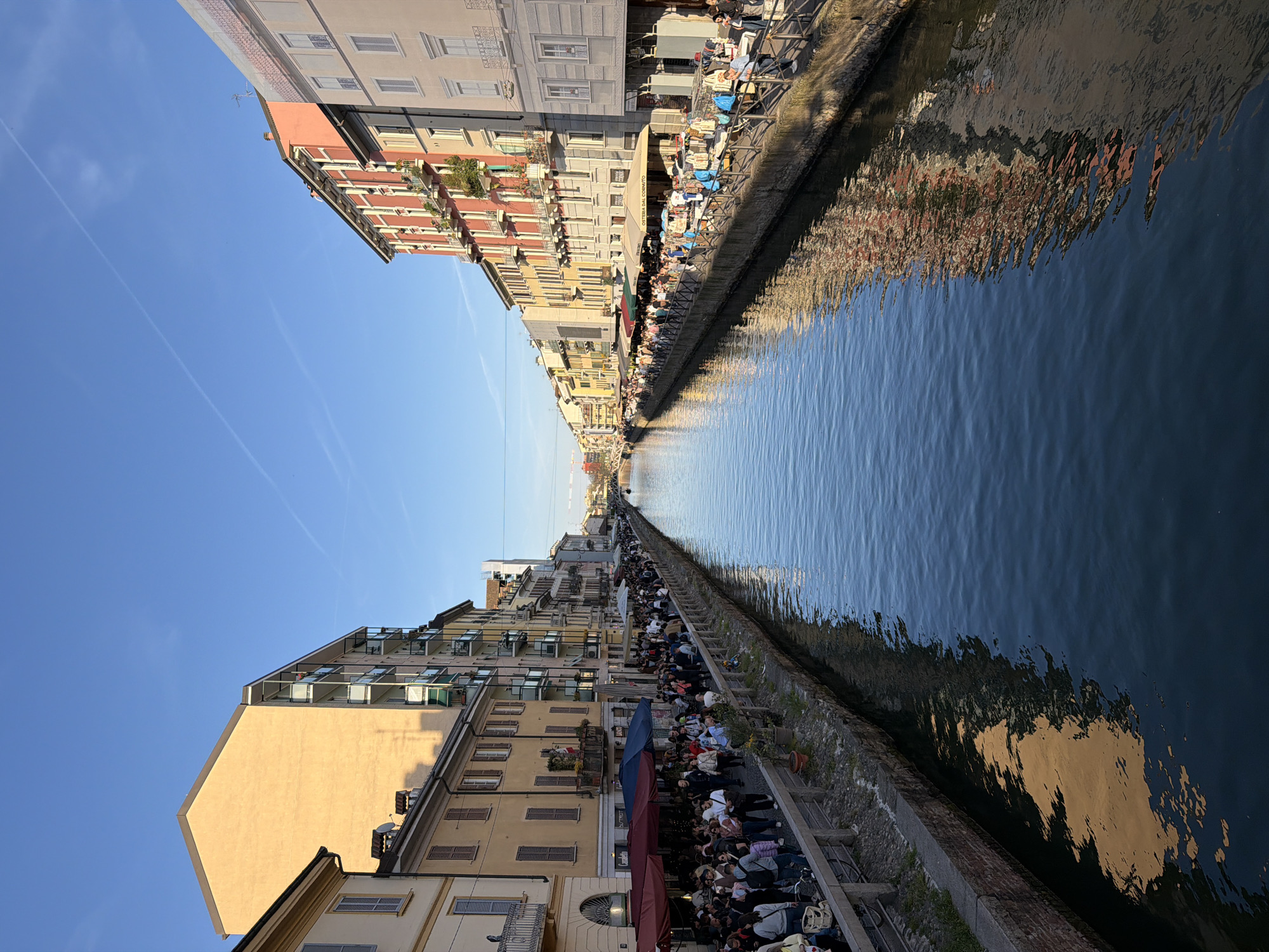 The image depicts the Naviglio Grande canal in Milan, Italy, lined with buildings and a bustling crowd. The buildings are varied in style, featuring balconies and windows. A large crowd of people congregates along the canal's edge, seemingly enjoying leisure activities at outdoor seating areas and on the sidewalks. The canal's water reflects the buildings and sky, showing slight ripples. The sky is blue with a few wispy clouds. The picture is taken during daylight hours.