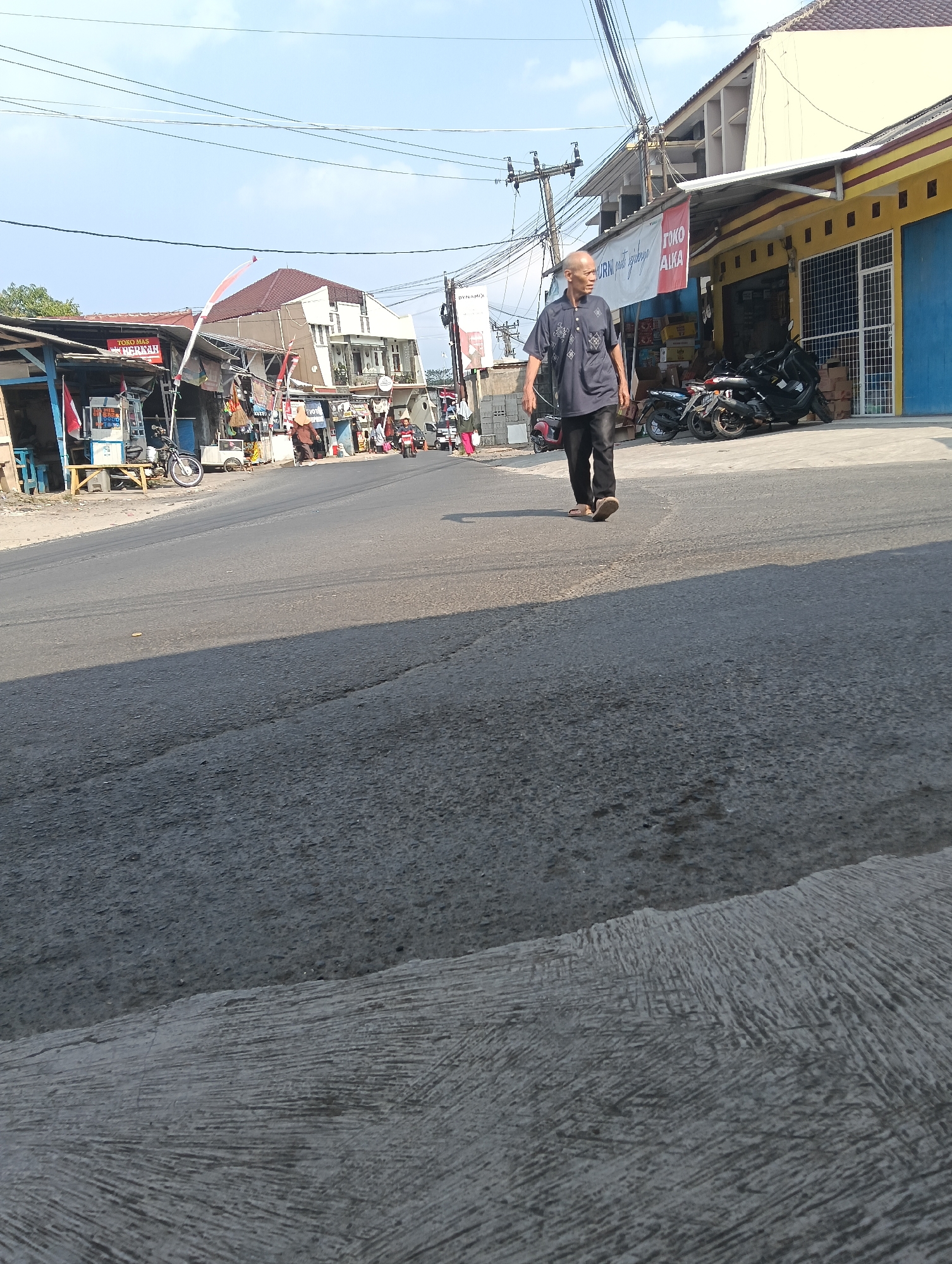 The image depicts a street scene in Puspahiang, Indonesia, under a clear sky. A bald, middle-aged man wearing a dark blue collared shirt with a patterned design and black pants is walking from left to right across the road. He is wearing sandals. To the left of the man are several storefronts with awnings. One storefront has a sign that reads "TOKO MAS BERKAH" (Gold Shop Blessing) and another sign in white with red text says "TOKO ALKA". Behind the man, there are parked motorcycles and a yellow building with a blue door. In the background, there are more buildings, including a multi-story white building with a brown roof. Power lines are visible overhead. The road is asphalt with visible cracks and a raised concrete curb on the right side of the image.
