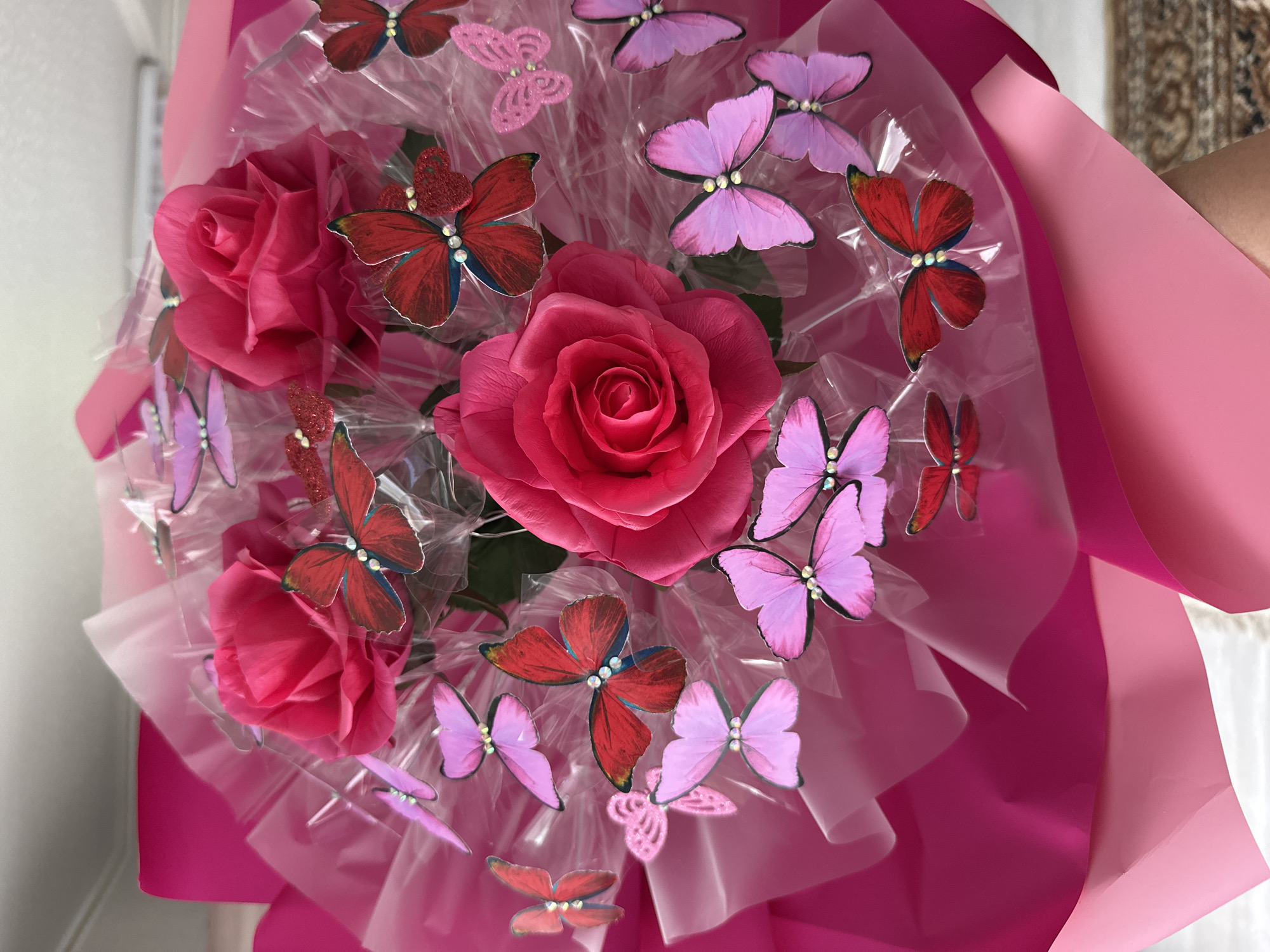 This is a close-up, eye-level, indoor photograph of a bouquet of pink roses adorned with numerous red and pink butterflies. The bouquet is wrapped in pink paper and clear plastic. The roses are fully bloomed, with rich pink petals. The butterflies are a mix of solid red with blue accents and solid pink, many featuring small sparkly embellishments on their bodies. The lighting is bright, creating a soft glow on the bouquet. The background is blurred, suggesting an indoor setting, possibly a home or shop, with a hint of a patterned rug visible at the bottom right. There is no visible indication of the location being in the city of Среднее Кечево, Russia.