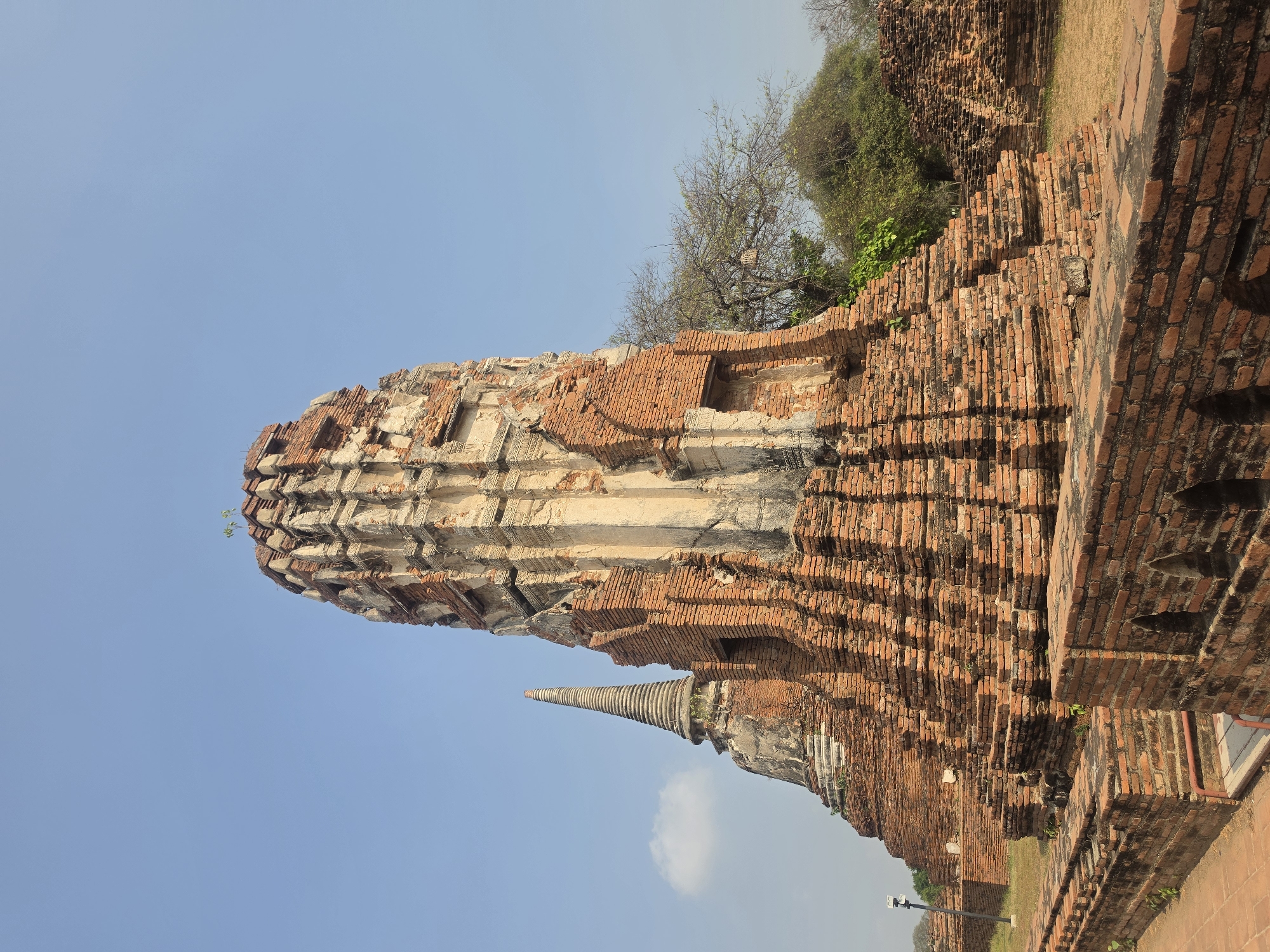 The image shows the ruins of Wat Yai Chaimongkol in Phra Nakhon Si Ayutthaya City Municipality, Thailand. The main structures are composed of red bricks and stone, with visible signs of weathering and damage. A large, partially crumbled chedi dominates the frame, with a smaller, spire-shaped chedi nearby. Some greenery grows on and around the ruins. The background includes trees and a clear, blue sky.