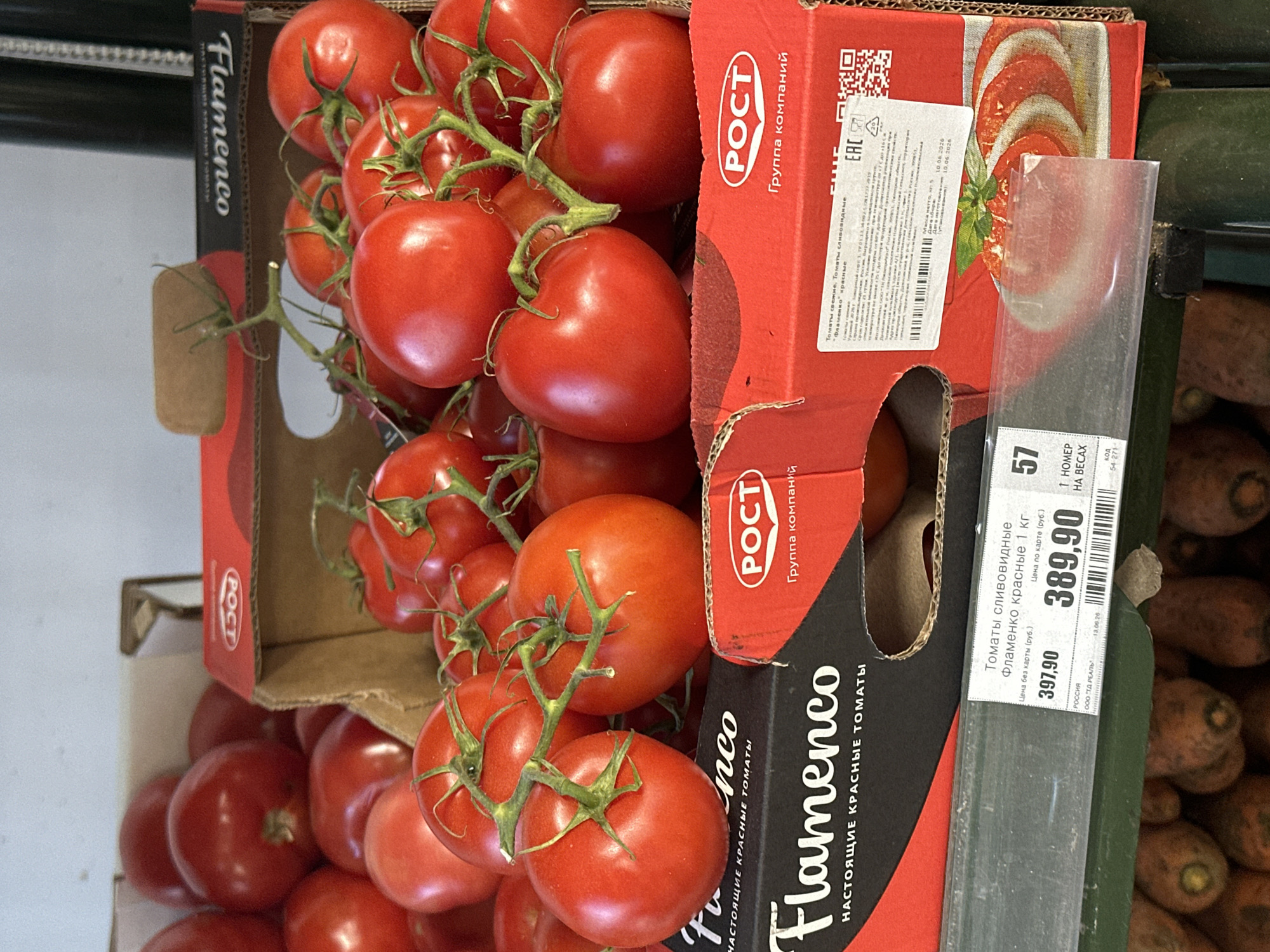 In Vsevolozhsk, Russia, a close-up shot captures a retail display of fresh tomatoes. The foreground is dominated by two cardboard boxes, one black and red, the other entirely red, both featuring the "Flamenco" brand name and "POCT" logos. These boxes are filled with ripe, red tomatoes, some still attached to their green stems. A price tag is affixed to the lower part of the display, showing prices in Russian Rubles for "Flamenco" tomatoes. In the background, more tomatoes are visible, along with glimpses of carrots in the lower portion of the frame. The lighting suggests an indoor retail environment, likely a supermarket or grocery store. No people are present in the image.