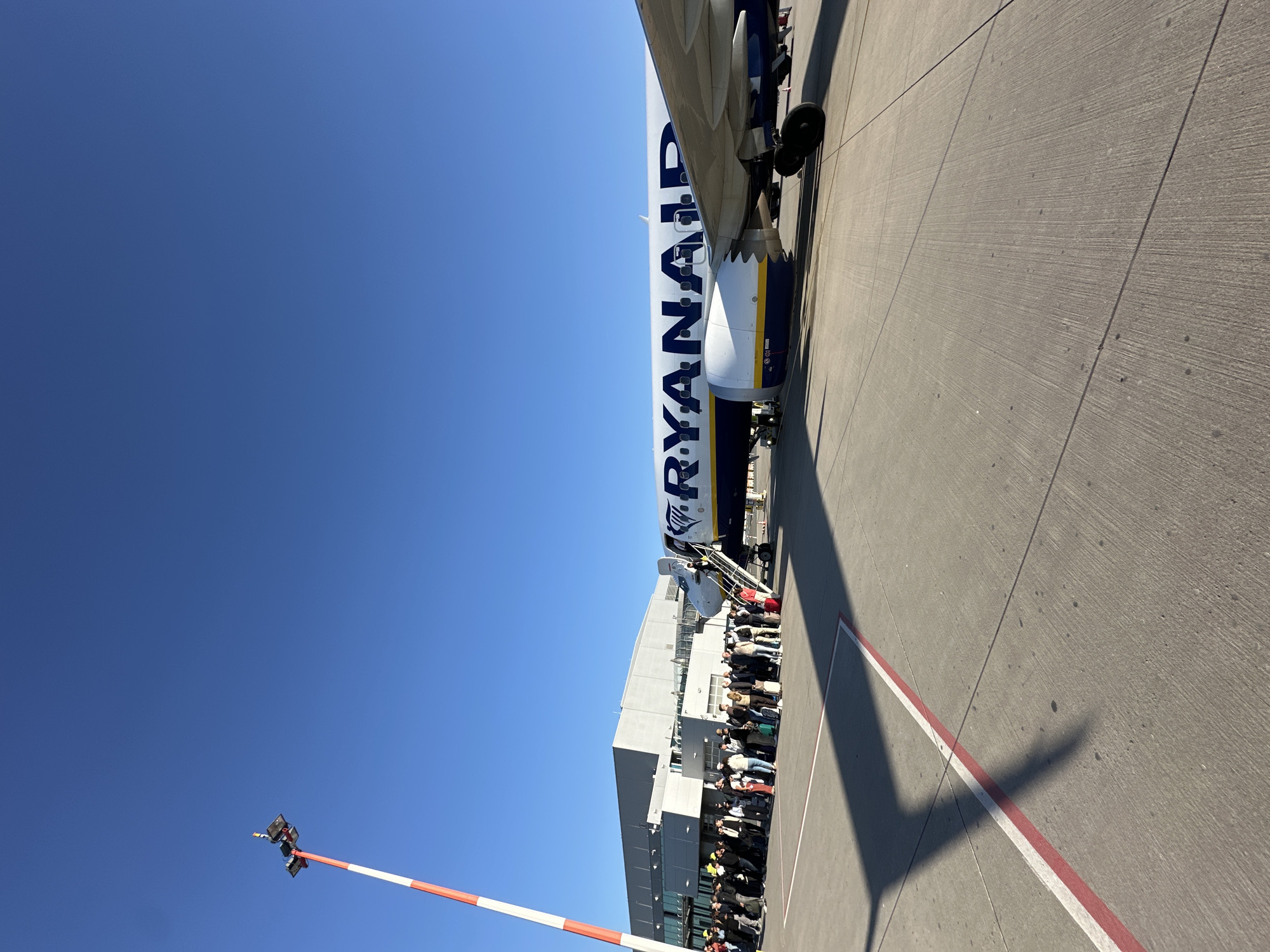 The image captures an outdoor scene at Weeze Airport, Germany, depicting a Ryanair airplane parked on the tarmac under a bright, clear blue sky. The aircraft's fuselage, emblazoned with the blue 'RYANAIR' text on a white background, dominates the upper right portion of the frame. A portable staircase is attached to the plane's door, leading to a visible entrance.
Below the plane, a large group of passengers is seen forming a queue on the tarmac, extending towards an airport building that runs parallel to the queue. The activity taking place is the boarding of the aircraft. Strong sunlight casts distinct shadows, including the outline of the airplane, onto the concrete ground which features red and white markings. This indicates a clear, sunny daytime. A red and white striped barrier pole is visible in the lower left corner against the vast blue sky.