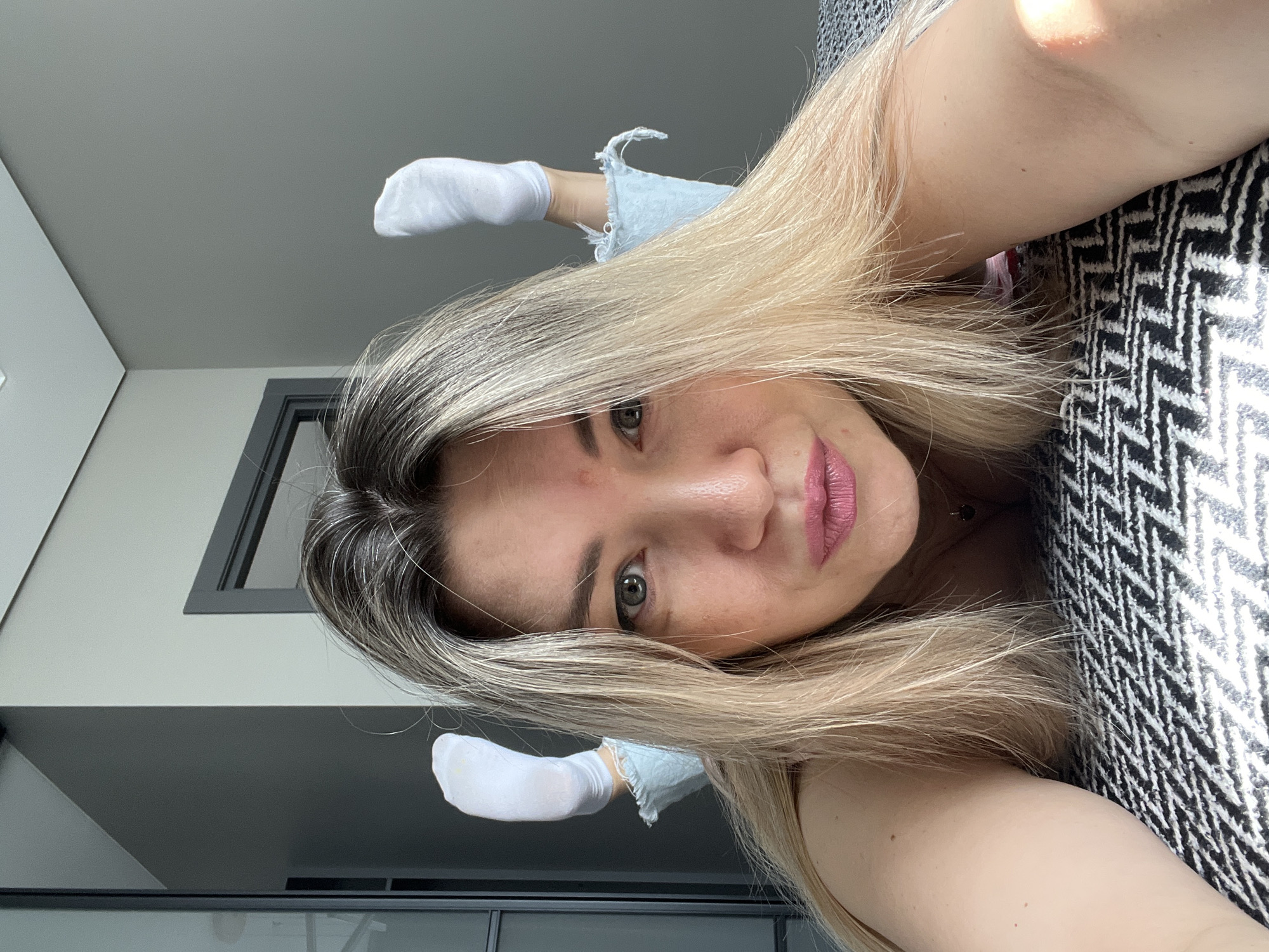 In a bedroom, a young woman with blonde hair and pink lipstick lies on a bed covered with a black and white patterned comforter. She is wearing light blue denim shorts with frayed edges and white socks. Her feet are raised in the air. The room is modern, with grey walls and a dark gray door frame visible in the background. The overall impression is casual and playful.