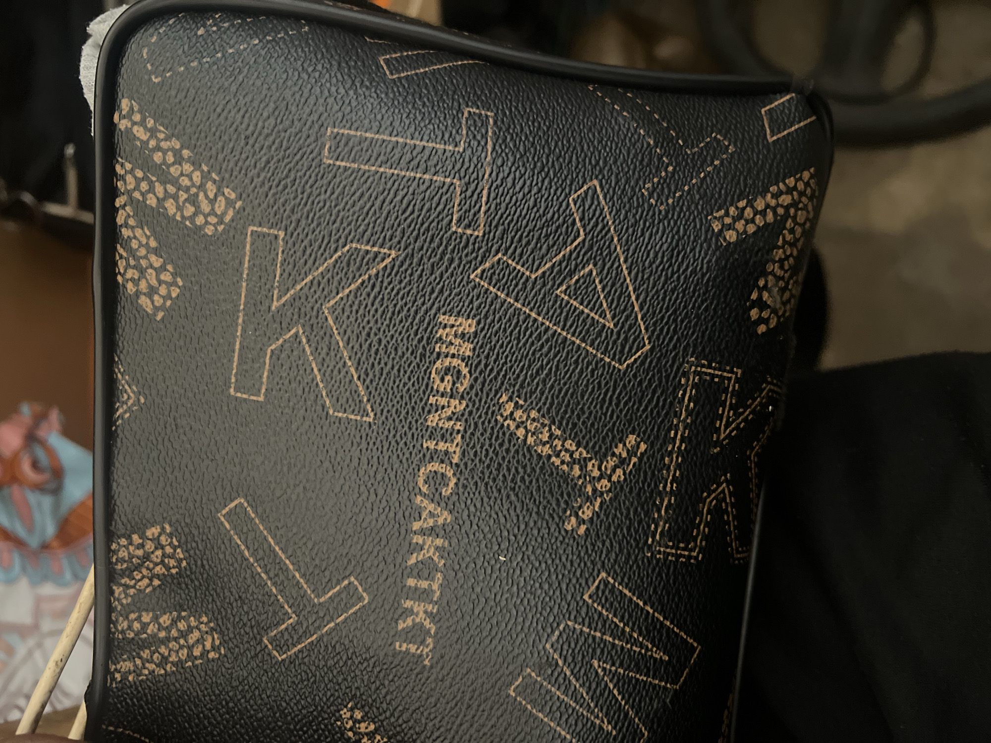 A close-up shot shows the textured surface of a black handbag with a repeating pattern of light brown letters and a speckled design. The word "MGNTCAKTKT" is visible in the center, printed in the light brown color. The bag appears to be made of faux leather. In the background, a colorful, blurred fabric is partially visible, along with a thin wooden stick and some white material. The overall scene is dark and the focus is on the handbag's surface and design. There is no identifiable context suggesting a location in Jalingo, Nigeria.
