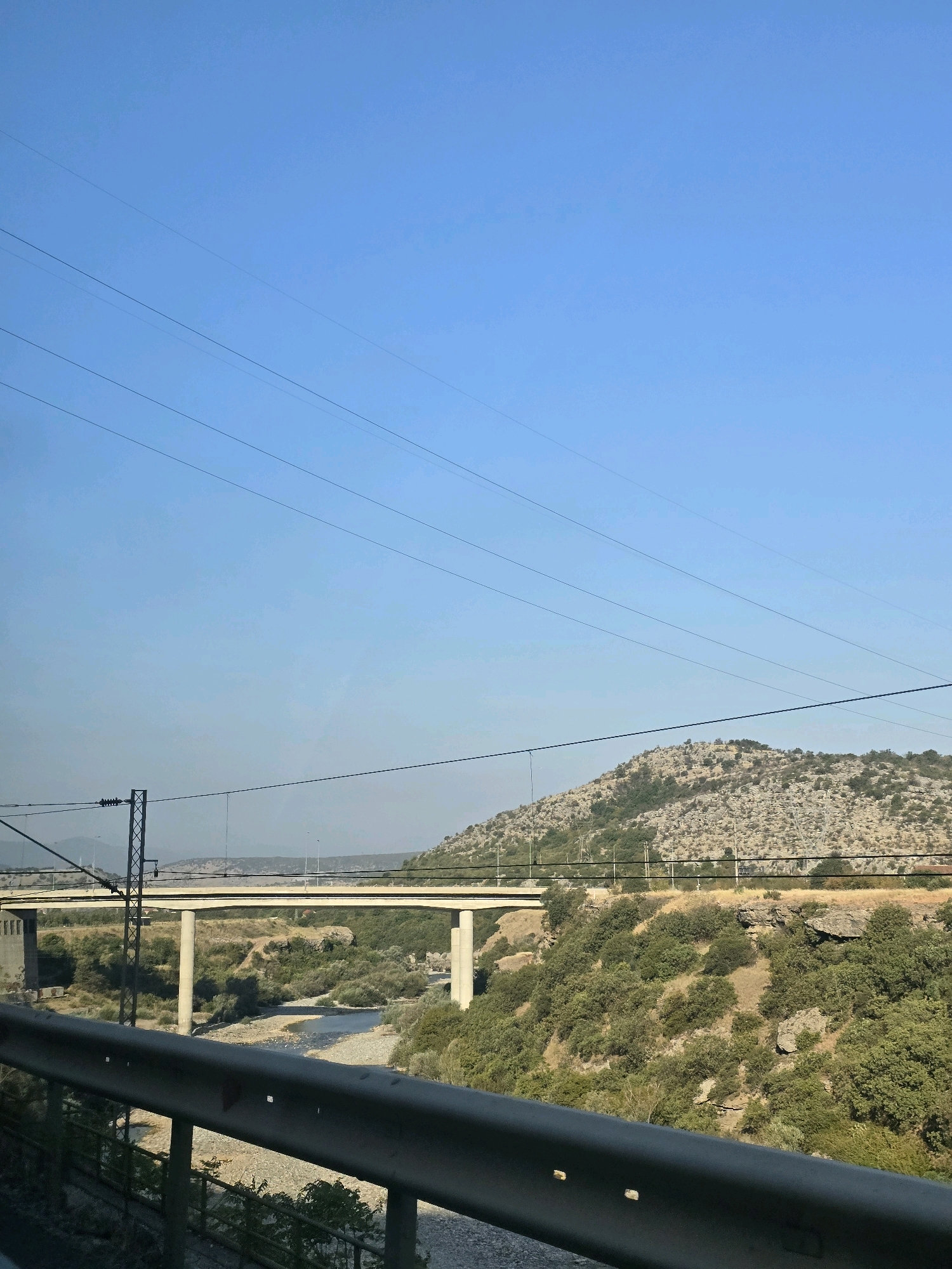 An exterior, medium shot captures a bright, clear sky with several power lines stretching horizontally across the frame. Below the sky, a wide, concrete bridge with multiple supporting pillars spans a shallow, rocky river. The bridge appears to be part of a highway or major roadway. The surrounding landscape is rugged and dry, with a barren, rocky hill rising in the background, sparsely covered in scrubby green vegetation. The foreground features a metal guardrail, suggesting the photo was taken from a vehicle. The scene is set in a rural or semi-rural area, possibly on the outskirts of a city. The lighting indicates it is daytime, with the sun high in the sky. This scene is likely a view from the highway that bypasses Podgorica, Montenegro, near the Morača river, a common route for travelers entering or leaving the city.