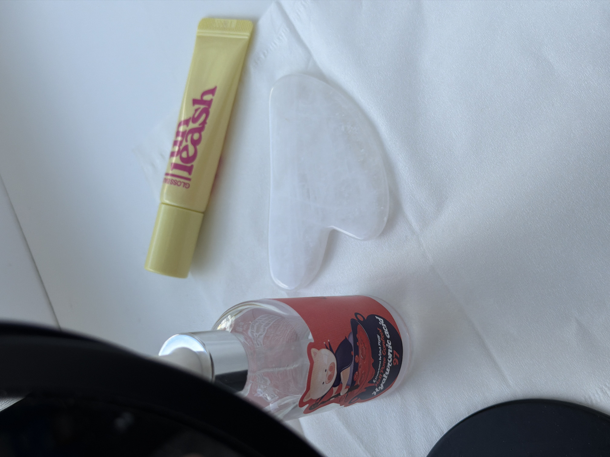 The image shows a close-up of a white textured surface, possibly a tablecloth or paper napkin. On this surface are three cosmetic items. To the left, a clear glass bottle with a silver pump dispenser contains a pink liquid and has a red label with a cartoon pig wearing a cape and holding a cauldron. Text on the label reads "Hyaluronic acid 97". In the center, a white, heart-shaped gua sha tool made of stone is visible. To the right, a yellow tube with pink lettering that reads "glossy leash" is also present. The overall lighting suggests an indoor setting, likely a bathroom or dressing area. There is no information in the image to suggest a specific location in Kyiv, Ukraine.