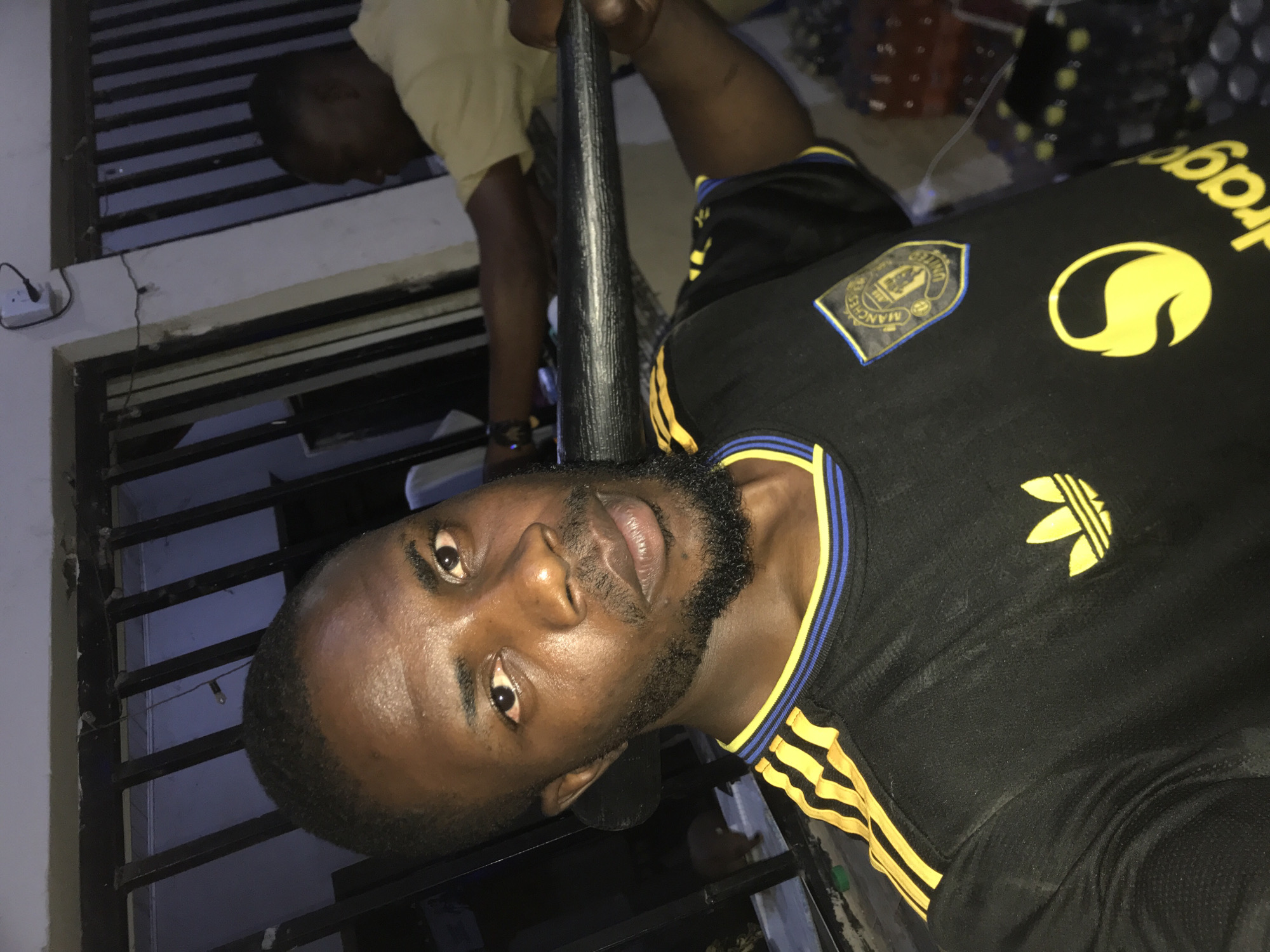 The image displays two males in an indoor or semi-enclosed environment, identified as being in New Stadium, Nigeria.
In the foreground, a Black male with short hair and a full beard directly faces the camera. He is wearing a black short-sleeved t-shirt with yellow Adidas stripes on the shoulders, a yellow Adidas Trefoil logo on the left chest, and a Manchester United Football Club crest patch on the right chest. A partial yellow "Snapdragon" logo is visible below the crest. He is holding a black, textured cylindrical object horizontally across his chest with both hands. His expression is neutral.
In the mid-ground, behind and to the right of the foreground subject, another out-of-focus Black male is partially visible. He wears a light yellow t-shirt and appears to be leaning forward, with his arms extended in an unspecified action.
The background consists of dark vertical metal bars, possibly part of a window grille or gate. A light-colored wall or column is present on the right, bearing a white electrical outlet with a black plug inserted. Several clear plastic bottles, some appearing to contain a yellow liquid, are stacked or arranged in the lower right background. The lighting is artificial and localized, illuminating the foreground subject prominently while the background remains darker.