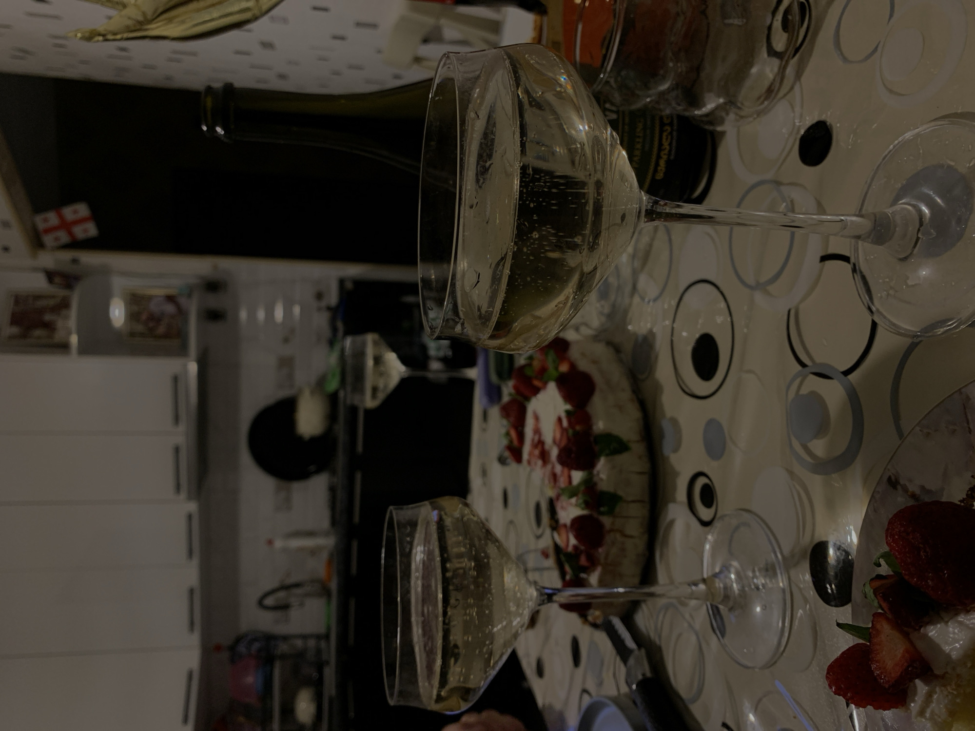 The image depicts a dimly lit indoor scene, likely a home kitchen in Rustavi, Georgia. In the foreground, two champagne coupes filled with a sparkling beverage are prominently displayed on a table covered with a patterned tablecloth. A partially eaten cake adorned with strawberries and greenery sits behind the glasses, suggesting a celebration. A bottle of sparkling wine is visible to the right, with a golden crown balloon partially obscuring the background. The kitchen cabinetry and appliances are visible in the background, with a small Georgian flag hanging from a doorway. The overall ambiance is one of informal festivity.