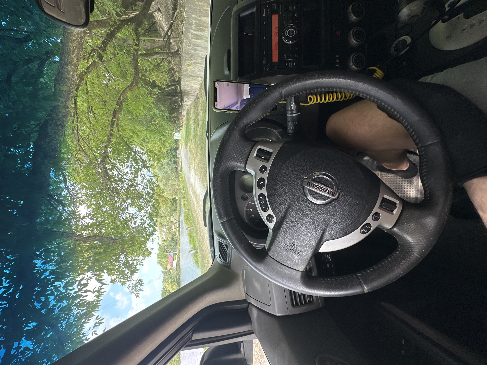 This image, taken from the interior of a Nissan, shows a view through the windshield towards a body of water and a tree-lined dirt road. The sun glints through the leaves of a large tree overhead, casting dappled light on the dashboard and steering wheel. A smartphone is mounted on the dashboard, displaying a pinkish screen. The car is parked on a dirt or gravel surface next to a low concrete wall. The presence of a lake or river, trees, and houses in the distance suggests a semi-rural or suburban setting. The specific location in Makiv, Ukraine, cannot be determined from the visual information alone.