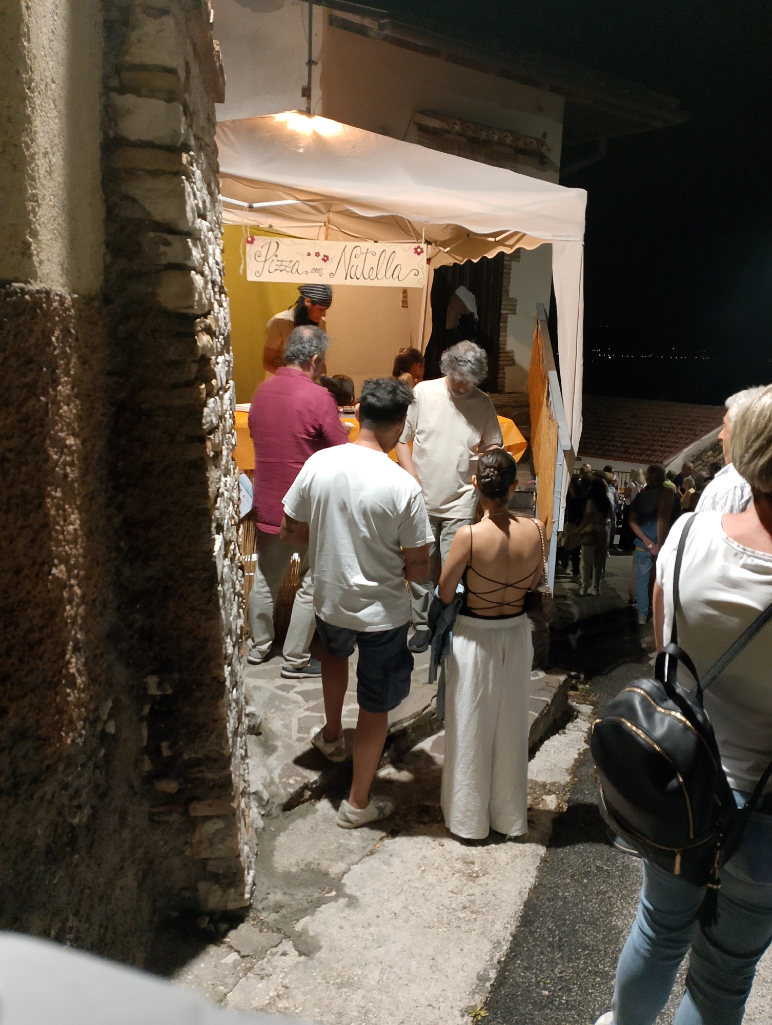 The image depicts a nighttime scene in Rivodutri, Italy, likely during a local festival or market. In the foreground, a stone wall runs along the left side of the frame. A white tent, illuminated from within, is set up, with a sign that reads "Pizza con Nutella" visible. Several people are gathered under or near the tent. To the left of the tent, a man in a red shirt is facing away from the camera, interacting with someone inside the tent. To his right, a man in a white t-shirt and blue shorts is also facing away from the camera. In front of them, a woman in a white outfit with a black laced top is standing. Further back, other individuals are visible, some appearing to be patrons of a food stall. The ground is paved with stones. In the background, a dark, hilly landscape is visible with distant lights, suggesting the presence of a town or city.