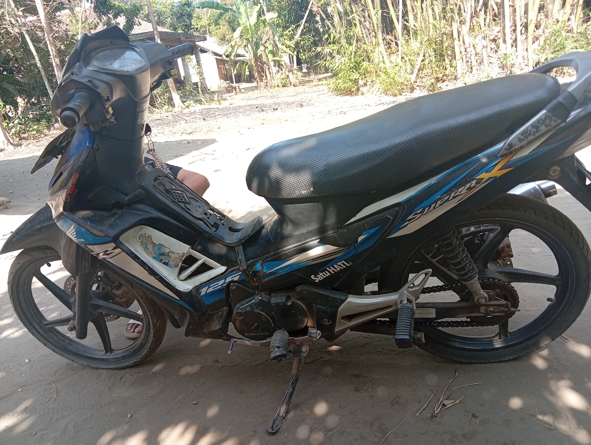 The image shows a black and blue Honda Supra X 125 motorcycle parked on a dirt ground. The motorcycle is leaning on its kickstand. The background shows vegetation, including trees and bamboo, and a building in the distance. The scene appears to be outdoors in a rural or semi-rural area. A person's legs, wearing flip-flops, are visible near the front wheel of the motorcycle, suggesting they may be the owner or attendant. The overall setting is consistent with a location in Miritpetikusan, Indonesia, which is a village in the Batangan district of the Pati Regency in Central Java.