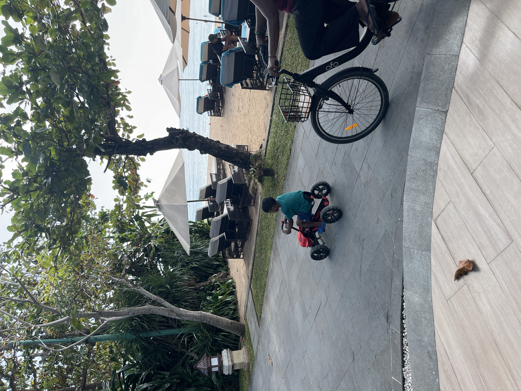 A boy rides a red pedal car on a tiled path in front of a beach in Kuta Selatan, Indonesia. The boy is wearing a teal t-shirt and blue shorts. To the right of the boy, a person on a black bicycle with a basket is also on the path. The beach has lounge chairs and umbrellas. Trees with green leaves are visible in the foreground. The sky is bright and overcast.