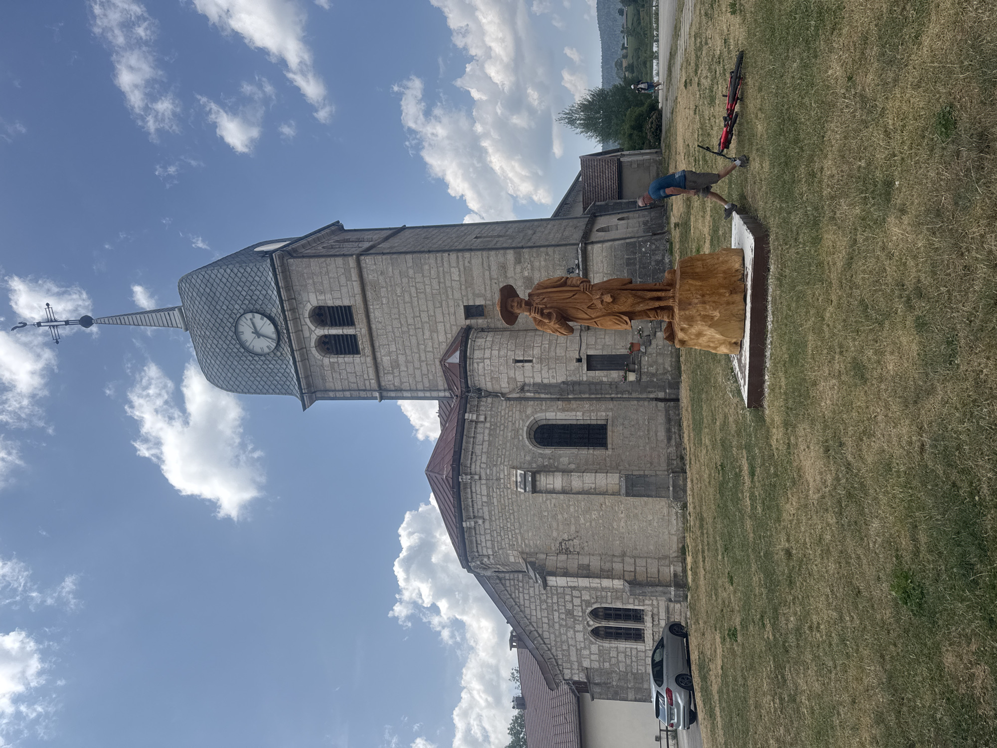 A full-shot image captures a scene in front of a stone church in Grande-Rivière Château, France. The church has a tall, square tower with a gray, shingled roof and a clock face on one side. A cross tops the tower. In the foreground, a large wooden sculpture depicts a man wearing a wide-brimmed hat, holding a stick, and standing next to a dog. The sculpture is placed on a wooden base. To the right of the sculpture, a man wearing a blue t-shirt and khaki shorts is walking past, with a red bicycle lying on its side in the grass nearby. The ground is covered in dry, yellowed grass. A silver car is parked to the left of the church. The sky is blue with scattered white clouds. In the background, a green hillside with trees is visible. Two other people are walking in the distance.