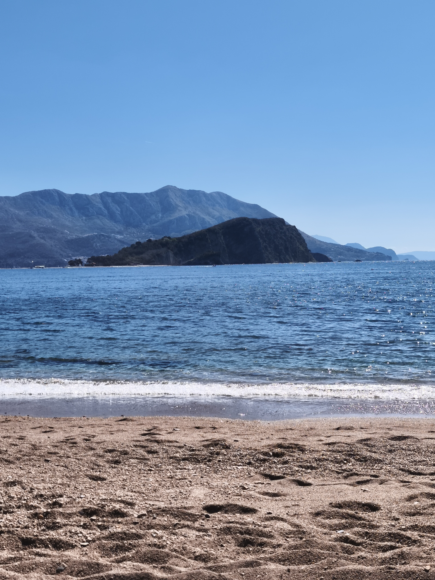 The image shows a beach scene in Budva, Montenegro. The foreground is a sandy beach with visible footprints. A narrow strip of water with small waves separates the beach from the calm, blue sea. In the background, a mountainous island is visible, with larger mountain ranges further inland. The sky is clear and blue. There are no people visible in the image.