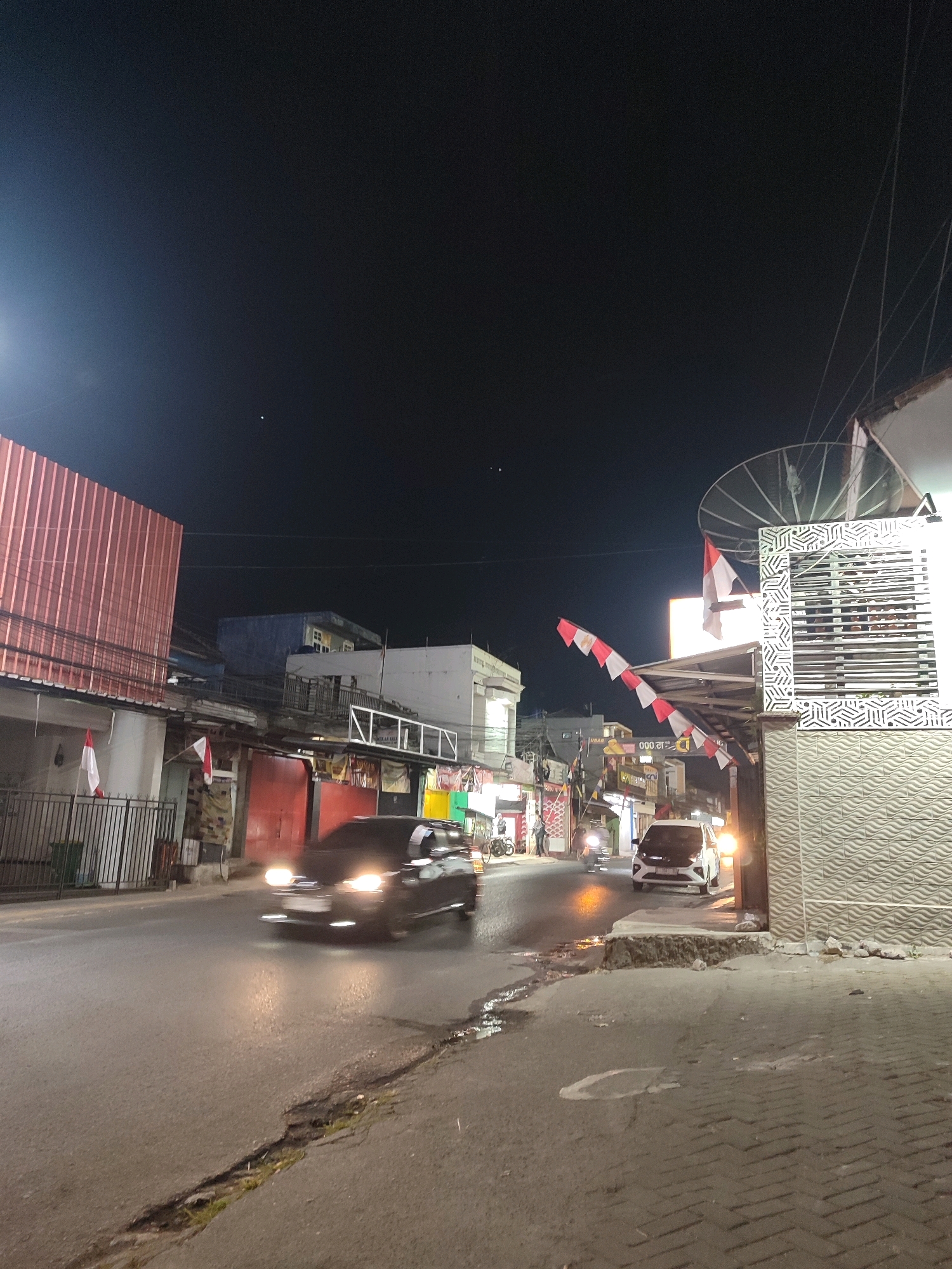 This nighttime street scene in Garut, Indonesia, depicts a modest commercial area. A dark-colored car, its headlights illuminated and blurred due to motion, is driving on the wet asphalt road. To the right, a white car is parked. The street is lined with buildings, one with a corrugated metal facade and another with a decorative screen. Red and white Indonesian flags are displayed on some buildings. The sky is dark, indicating night, and a satellite dish is visible on one of the structures. The sidewalk is paved with bricks and shows signs of wear and tear.