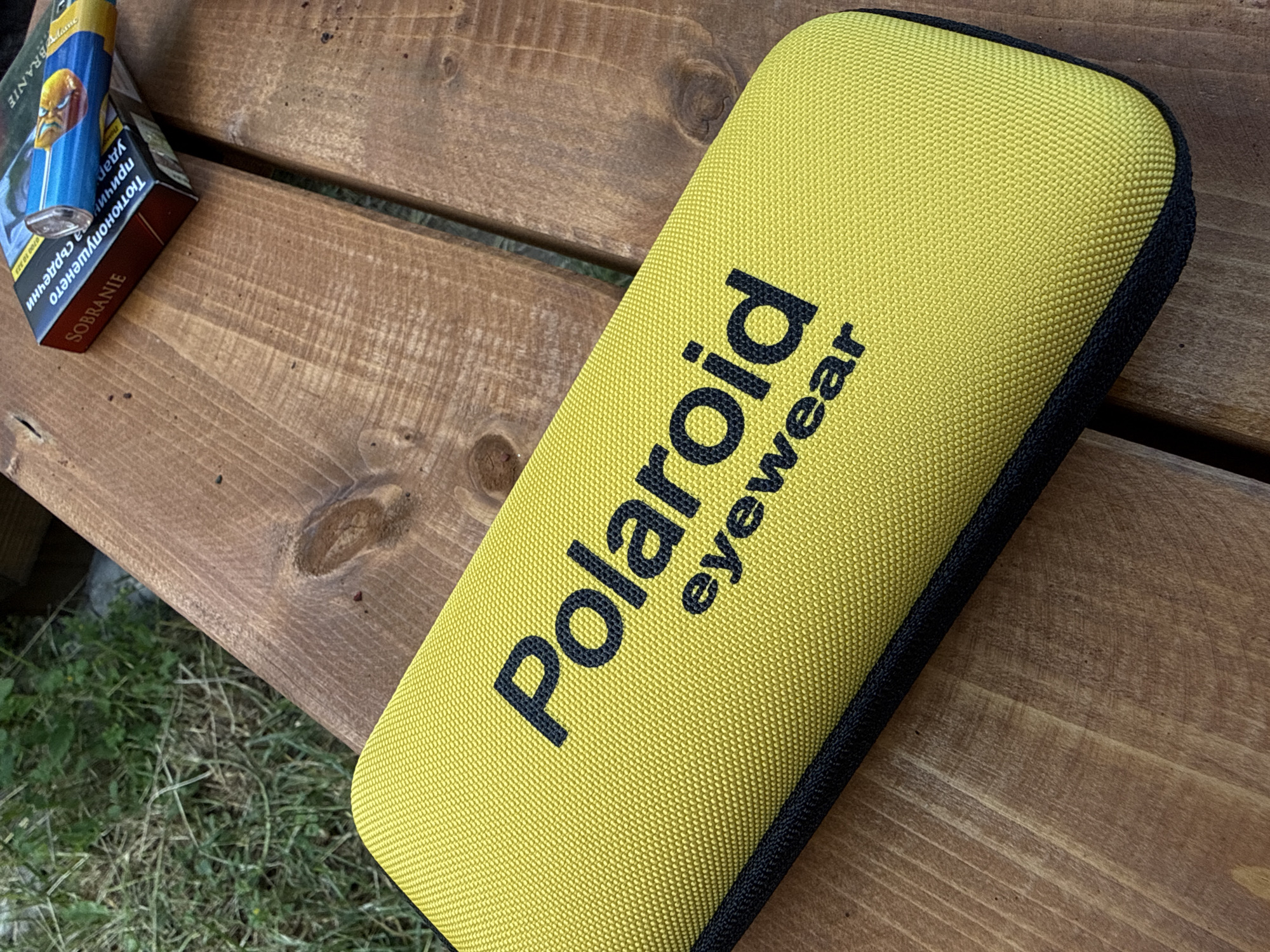 A close-up, high-angle outdoor shot shows a bright yellow eyeglass case with black lettering that reads "Polaroid eyewear." The case rests on a weathered wooden table, with a pack of Sobranie cigarettes partially visible in the upper right corner. In the background, out of focus, is green grass and dry vegetation. The lighting suggests it is daytime. The image does not depict any specific location in Zgorigrad, Bulgaria, or any people.